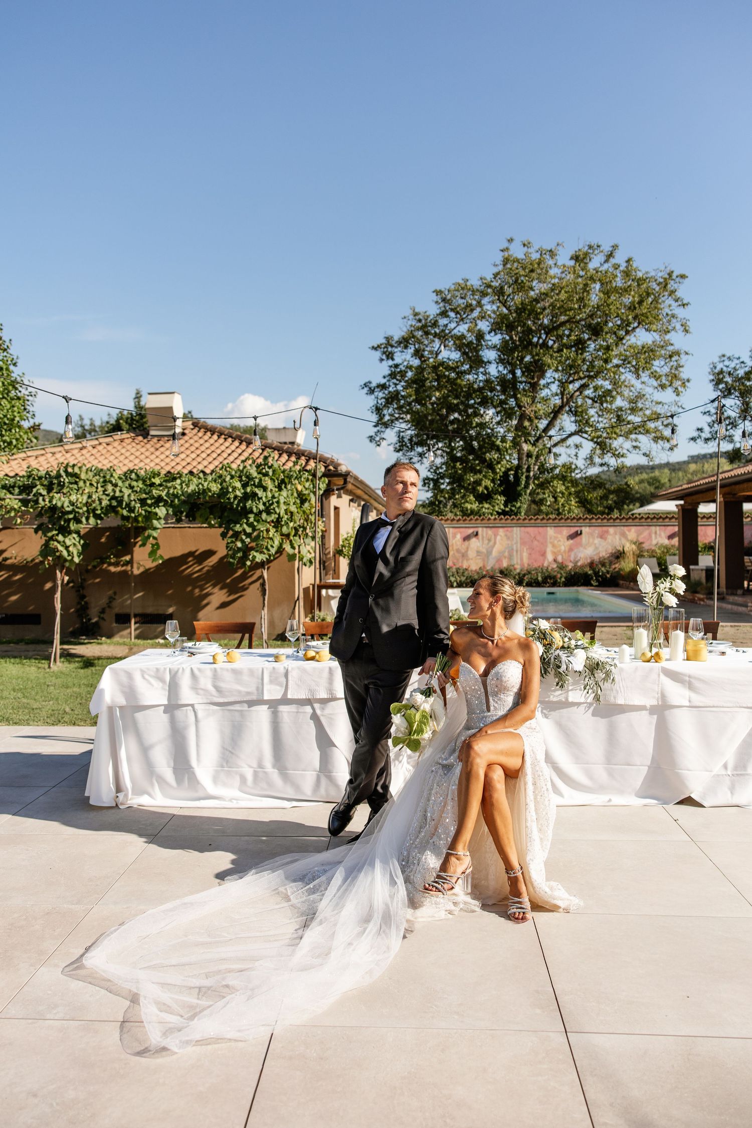 Elegant bride in lace gown sits beside groom in black suit at outdoor wedding reception venue with lush greenery.