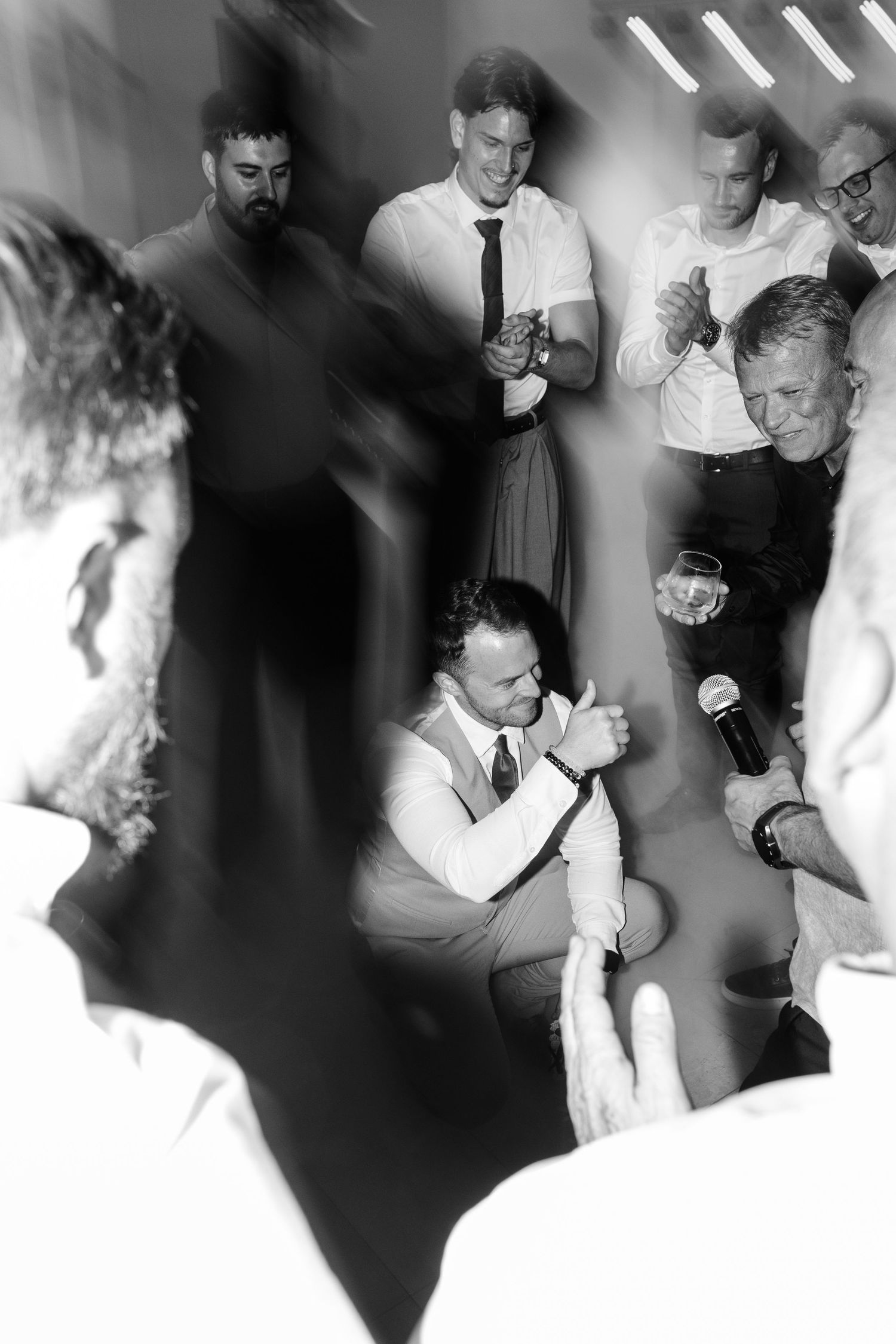 Black and white photo of lively wedding reception with groom kneeling amid cheering guests.