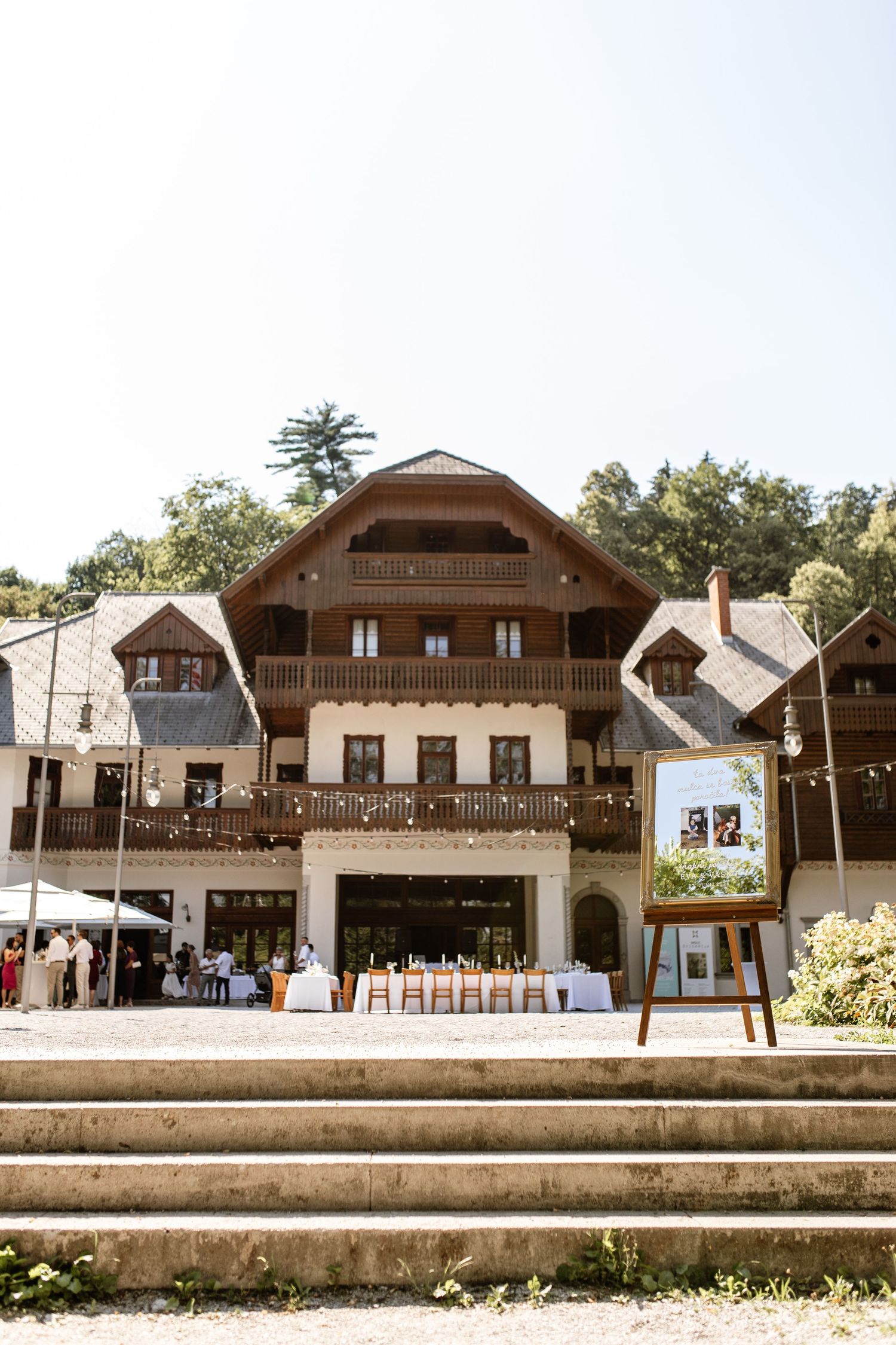 Elegant multi-story chalet venue with wooden balconies, string lights, and outdoor wedding reception setup on sunny day.