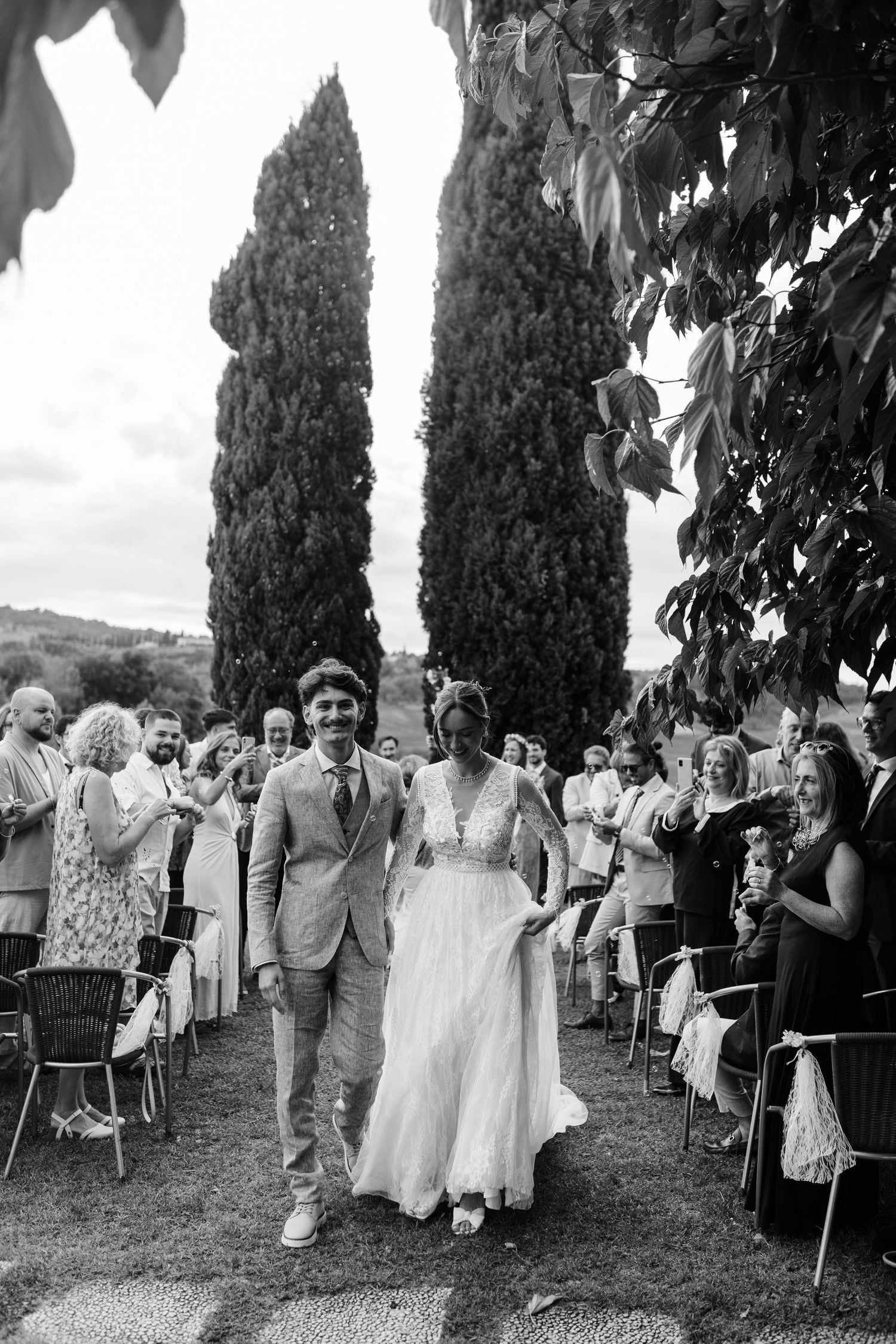 Black and white photo of a smiling couple walking down the aisle at an outdoor wedding ceremony with cypress trees.