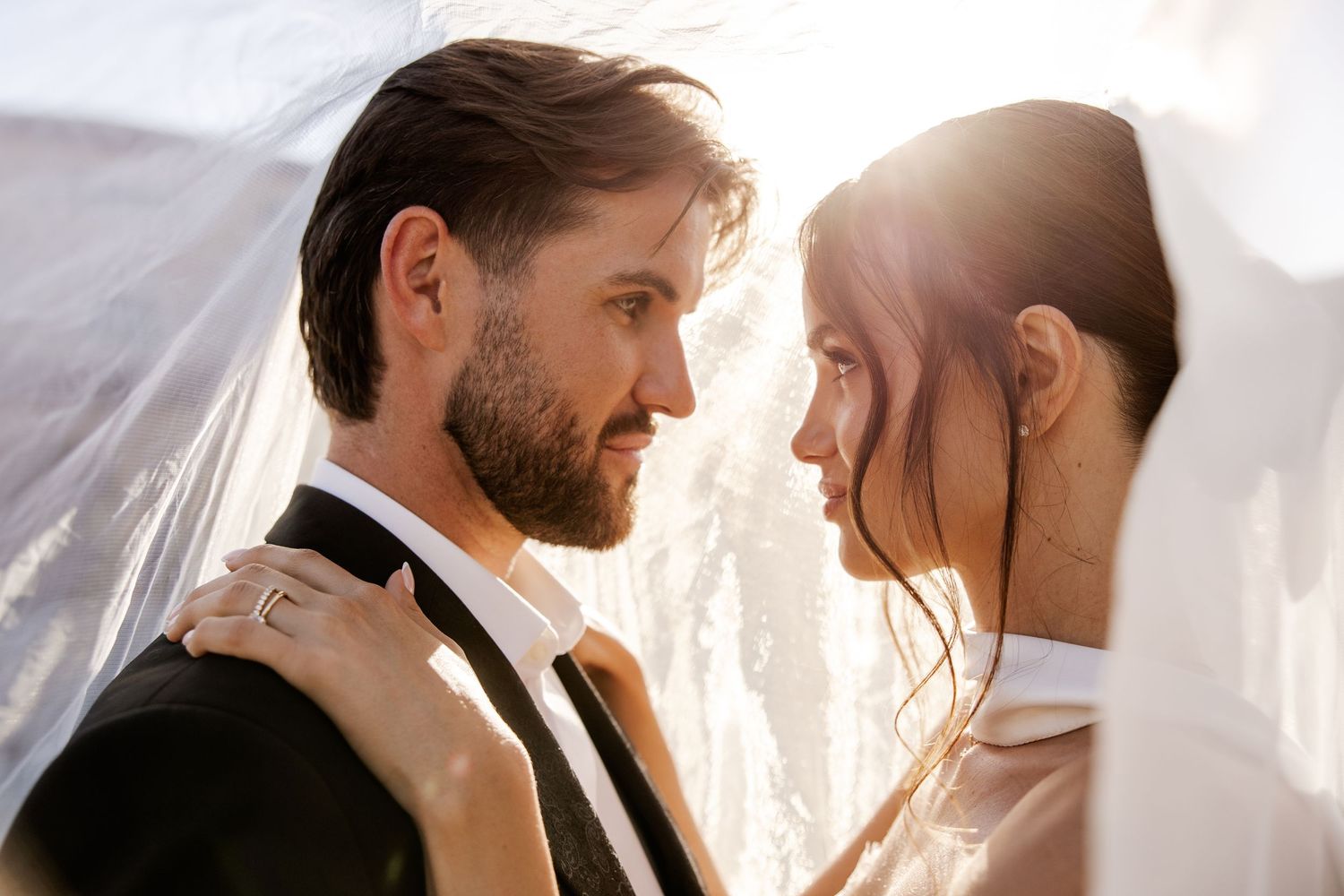 Romantic wedding couple sharing an intimate moment under a bridal veil in golden sunlight.