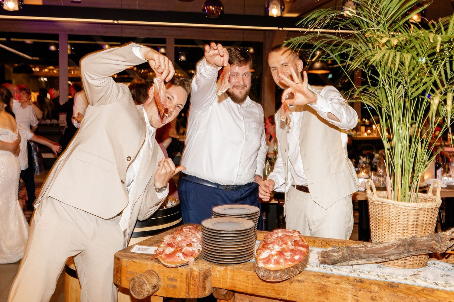 Three men in white suits making hand gestures at a wedding reception with charcuterie display.