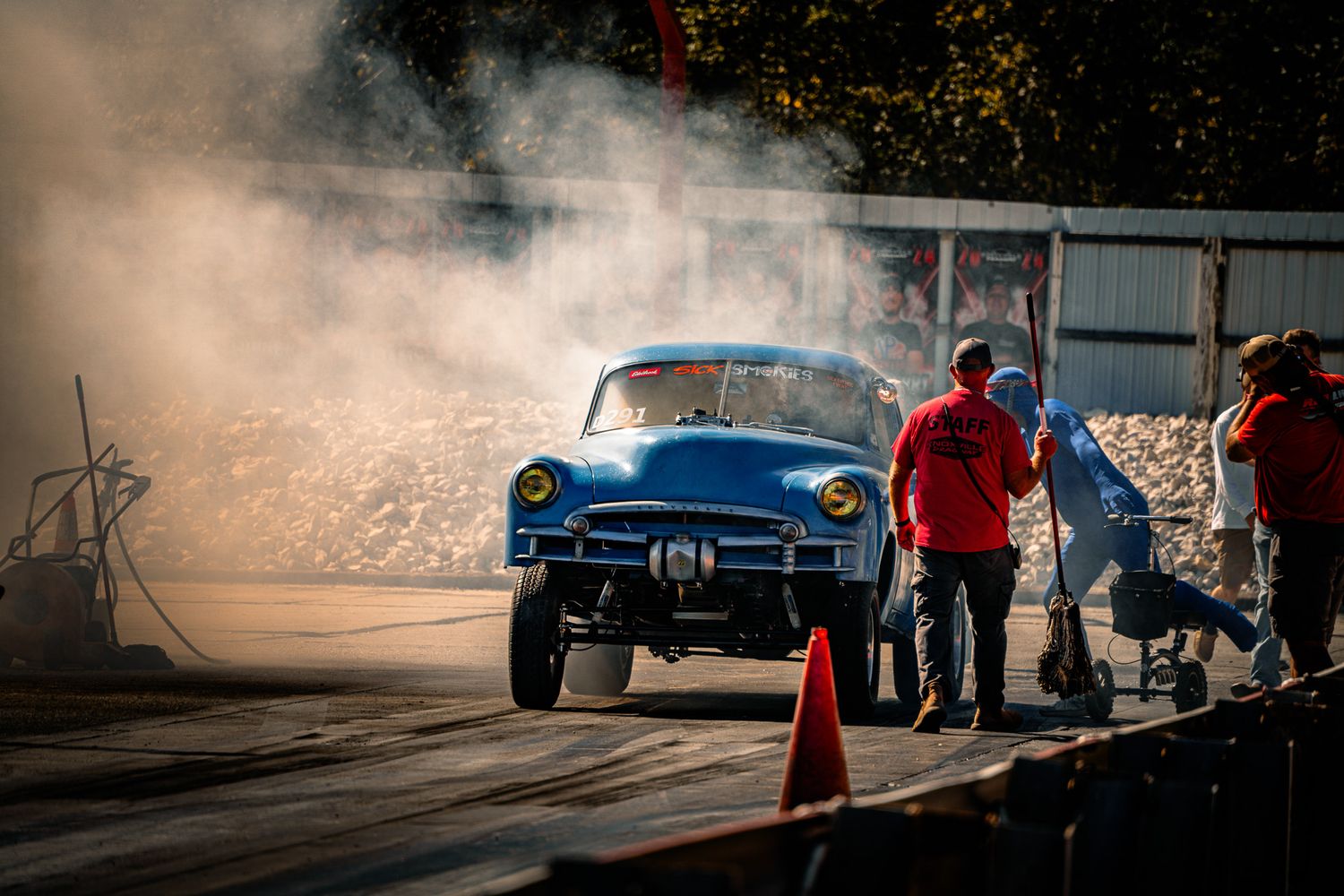Blue vintage gasser car performing burnout with smoke on drag strip surrounded by crew members in red. Sick Smokies