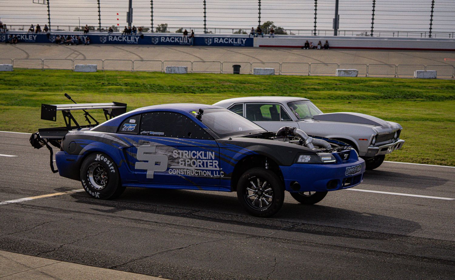 Two drag racing cars competing on a professional racetrack, including a blue Mustang with spoiler. Street Car Takeover