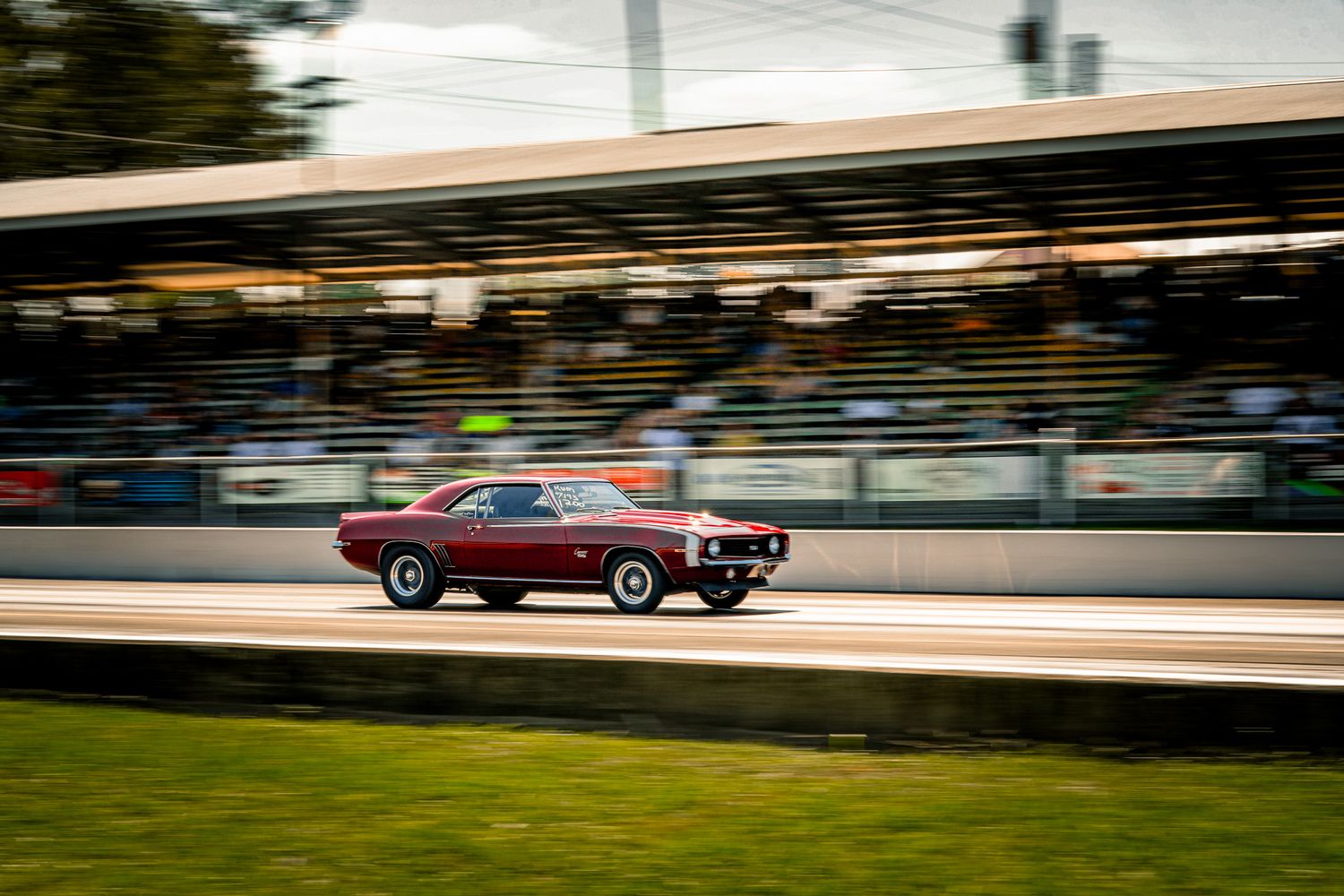Dark red classic muscle car racing at high speed on drag strip with grandstands. Holley LS Fest