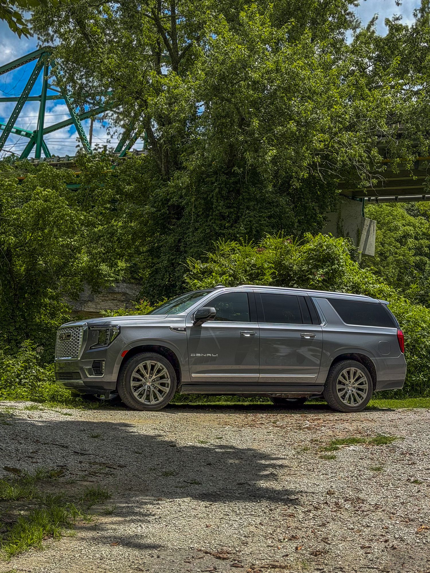 Gray GMC Yukon XL full-size luxury SUV parked on gravel near lush green trees.