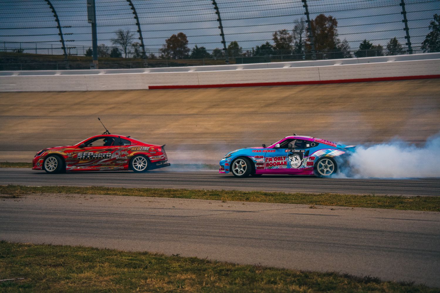Two drift cars producing smoke on a race track during a tandem drifting competition. Slyde Factory