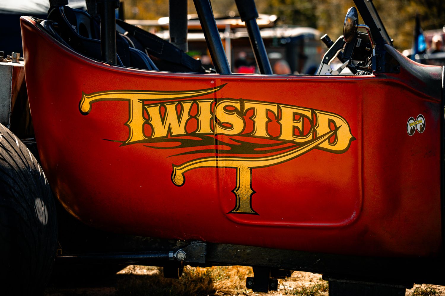 Close-up of red dragster body panel with gold Twisted T logo lettering at an outdoor racing event. Sick Smokies