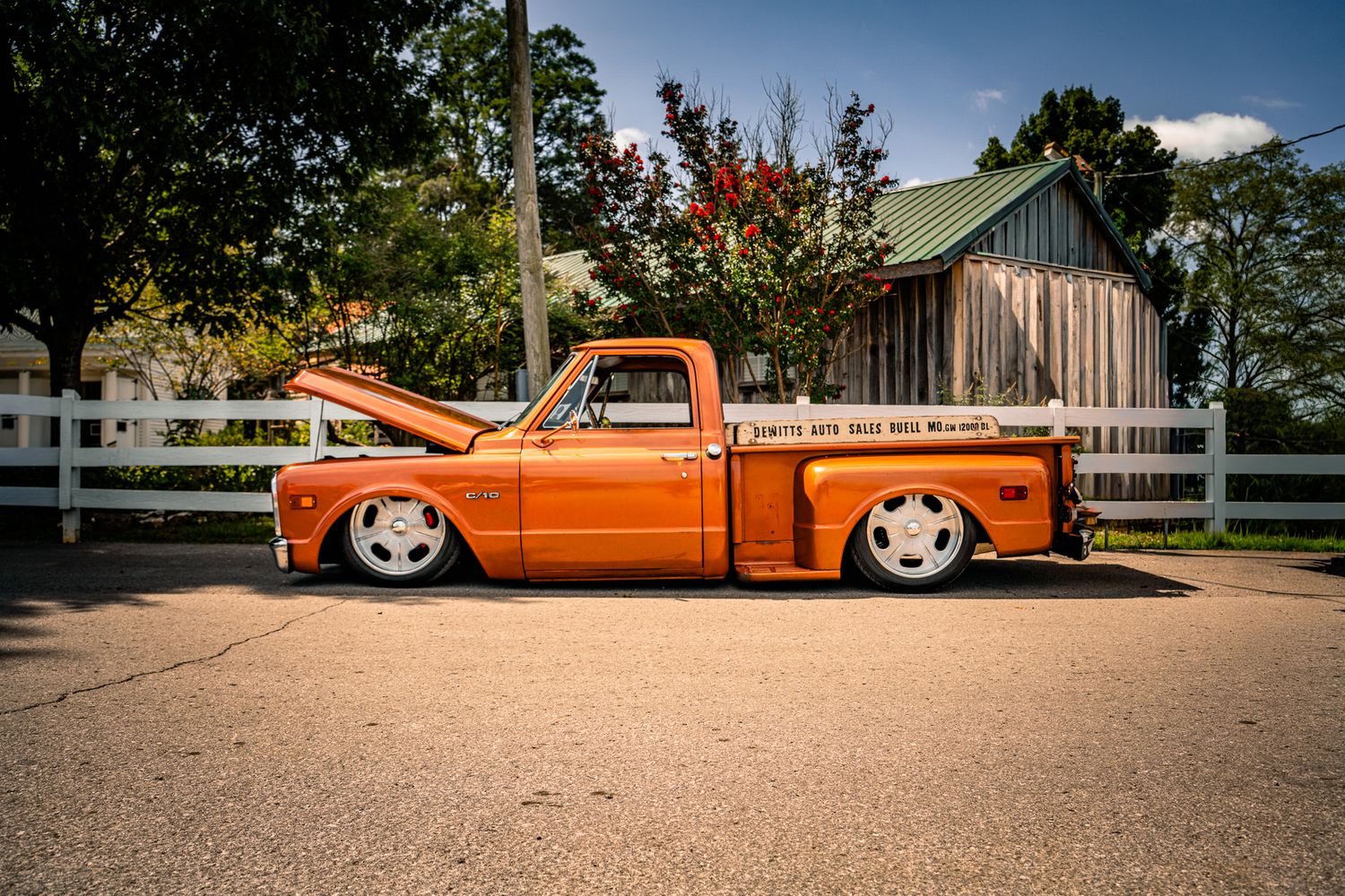 Custom orange lowrider pickup truck with hood open parked beside white fence and barn. Southeastern Truck Nationals