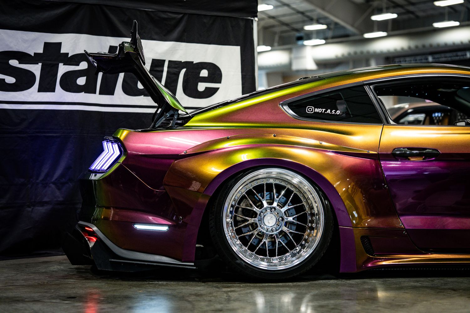 Custom Ford Mustang with iridescent purple-gold wrap and wide-body kit on display indoors. Slammednuff