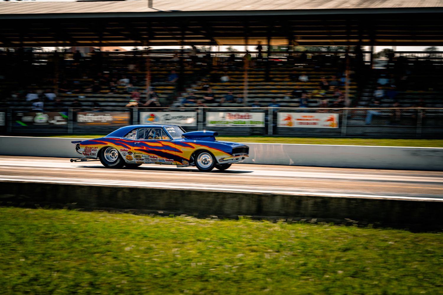 Flame-painted classic muscle car racing down drag strip at dusk with grandstands visible in background. Holley LS Fest