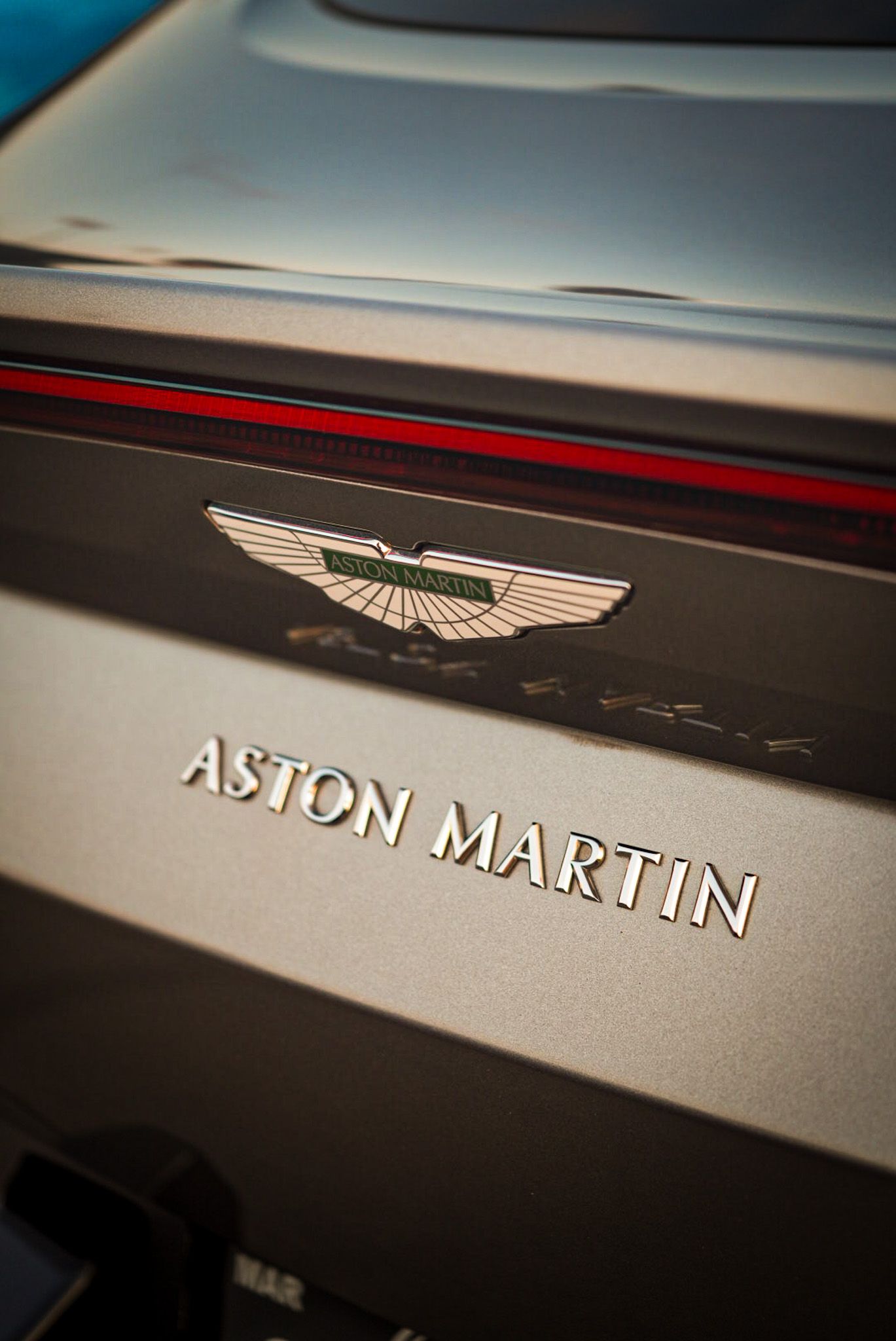 Close-up of Aston Martin logo and badge on a sleek silver luxury sports car exterior. Cars & Christ