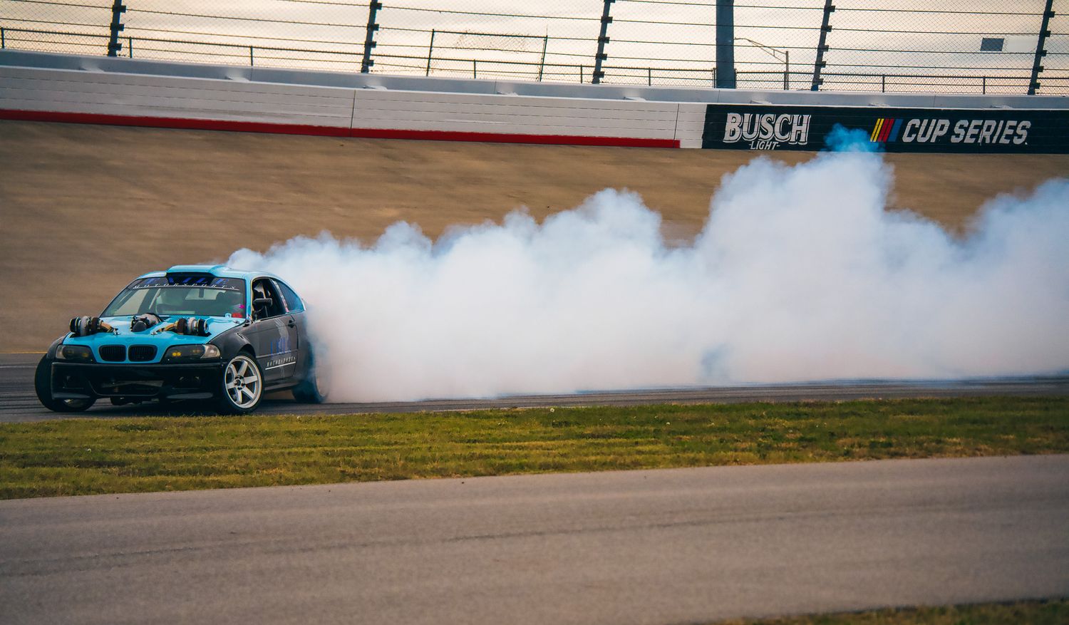 BMW sedan performing dramatic drift with massive tire smoke at Busch Cup Series track. Slyde Factory