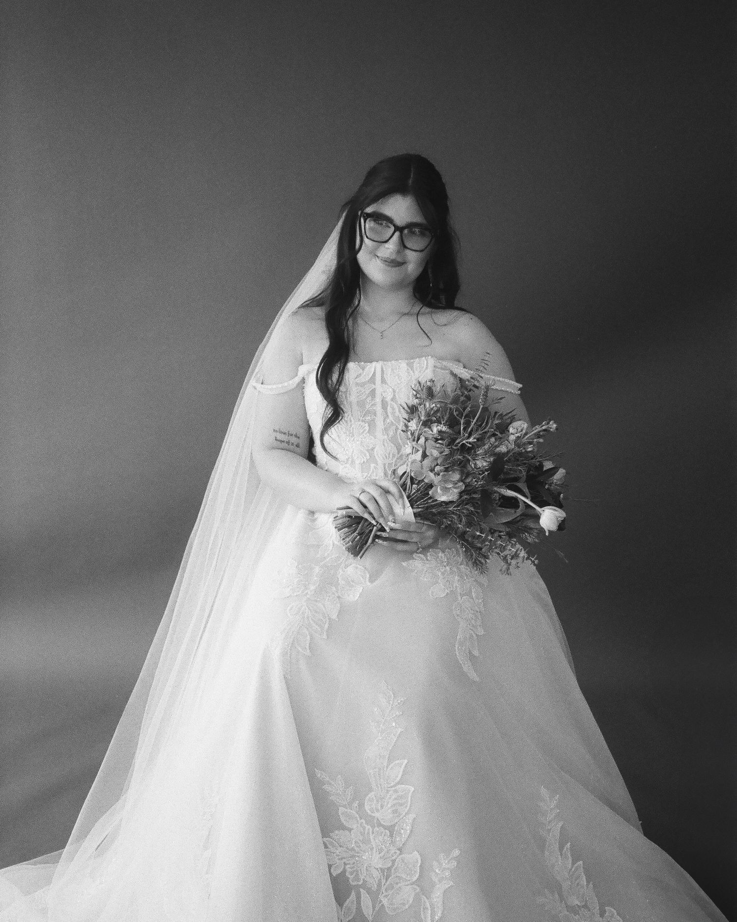 Black and white portrait of bride in off-shoulder gown with veil and glasses holding bouquet.