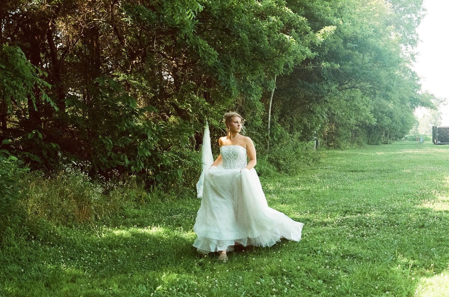 Bride in strapless white wedding gown walking gracefully across a lush green lawn surrounded by tall trees.