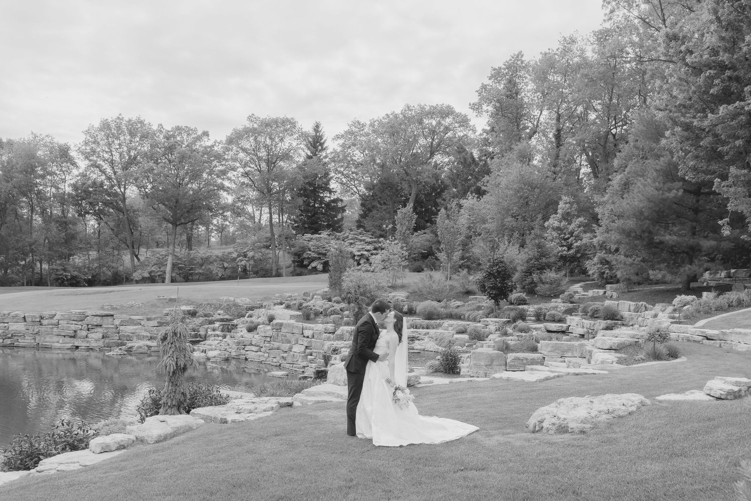 Black and white wedding portrait of a couple standing near a serene pond in a garden.