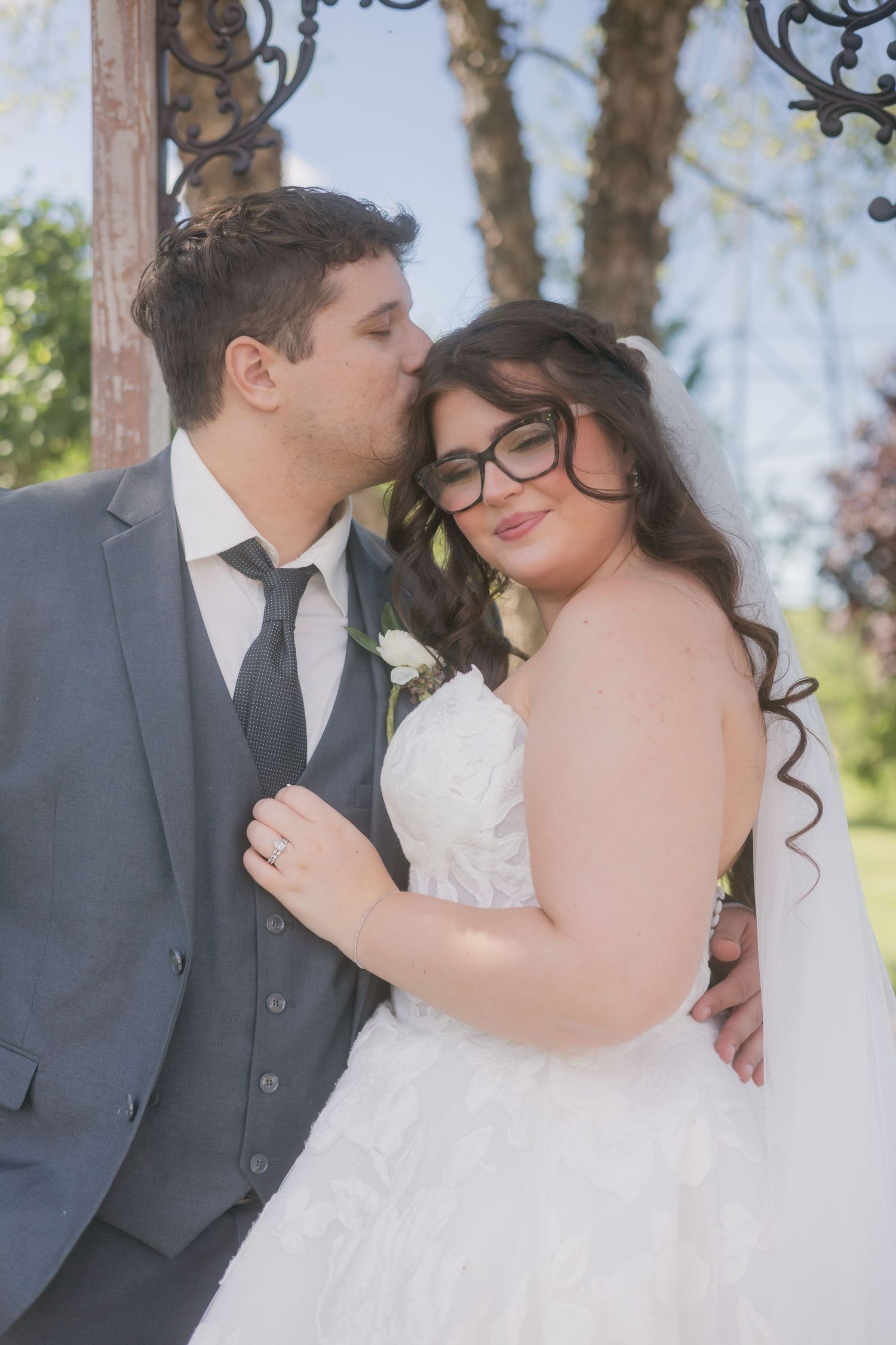Groom in gray suit kisses bride's cheek as she smiles wearing glasses and lace wedding gown.