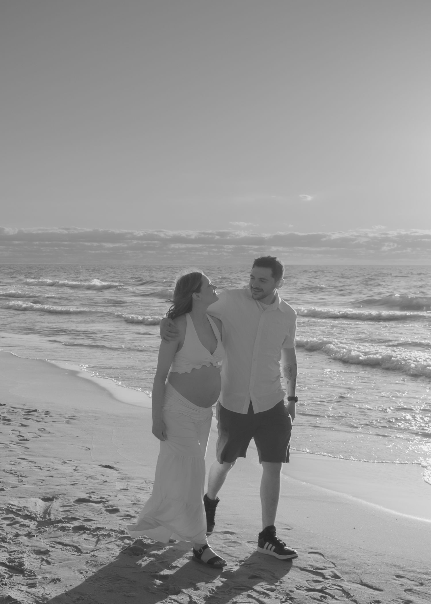Black and white photo of couple walking hand in hand along a sunny ocean beach.