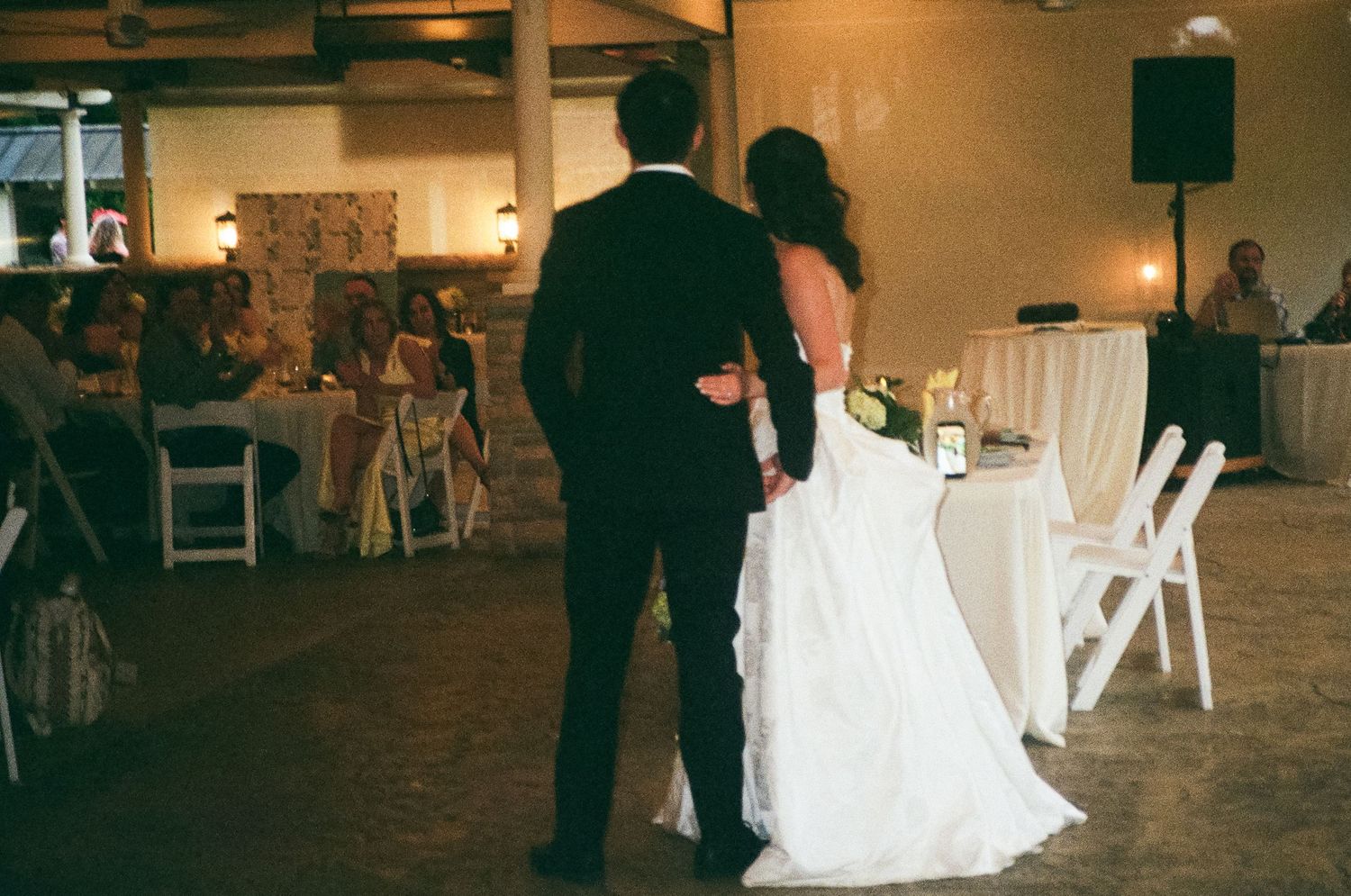 Bride and groom stand hand in hand facing wedding reception guests in warmly lit venue hall.