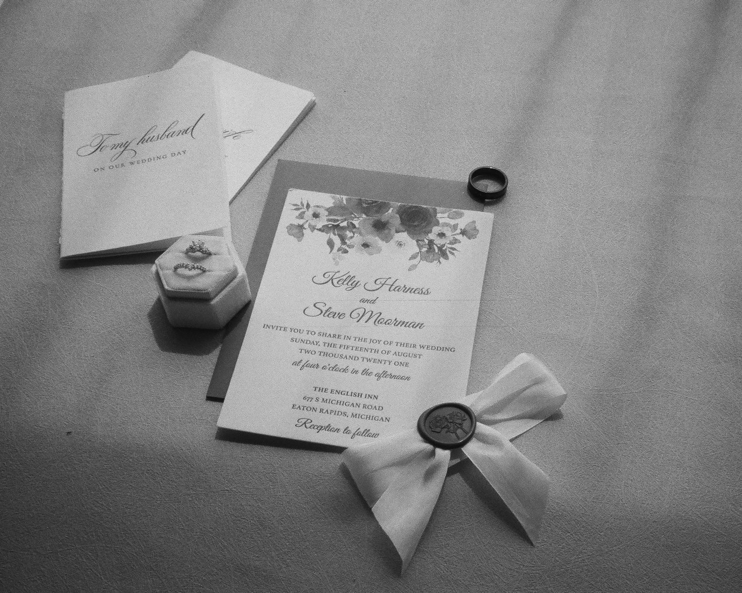 Black and white wedding invitation suite with floral details, wax seal, ribbon bow, ring box, and wedding band on soft fabric.