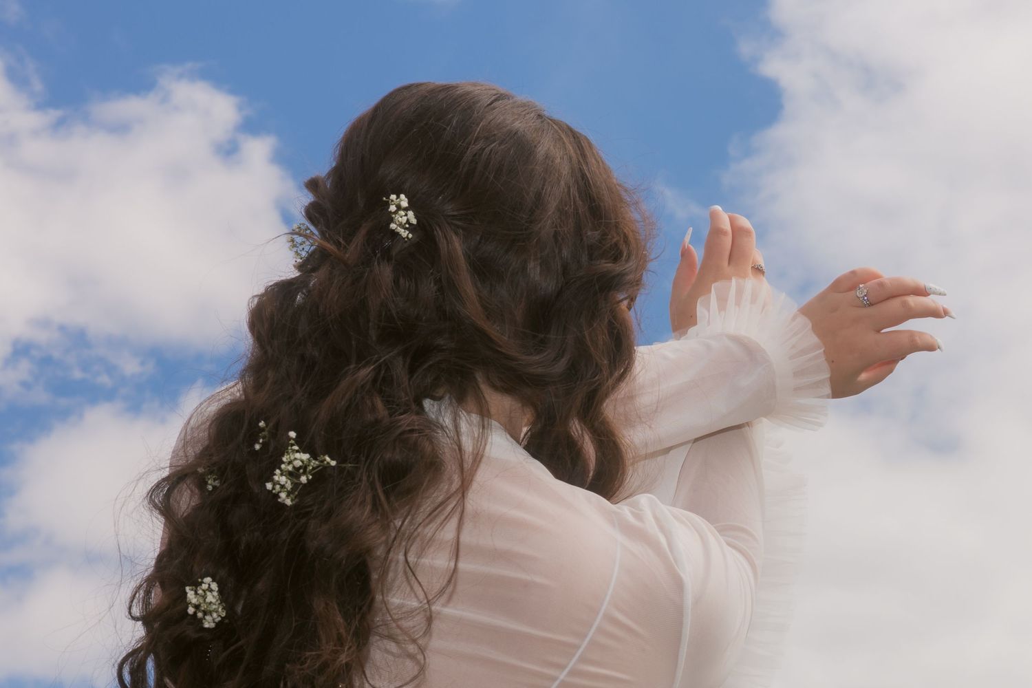 Bride with long dark curly hair adorned with small white flowers, wearing white dress against blue sky.