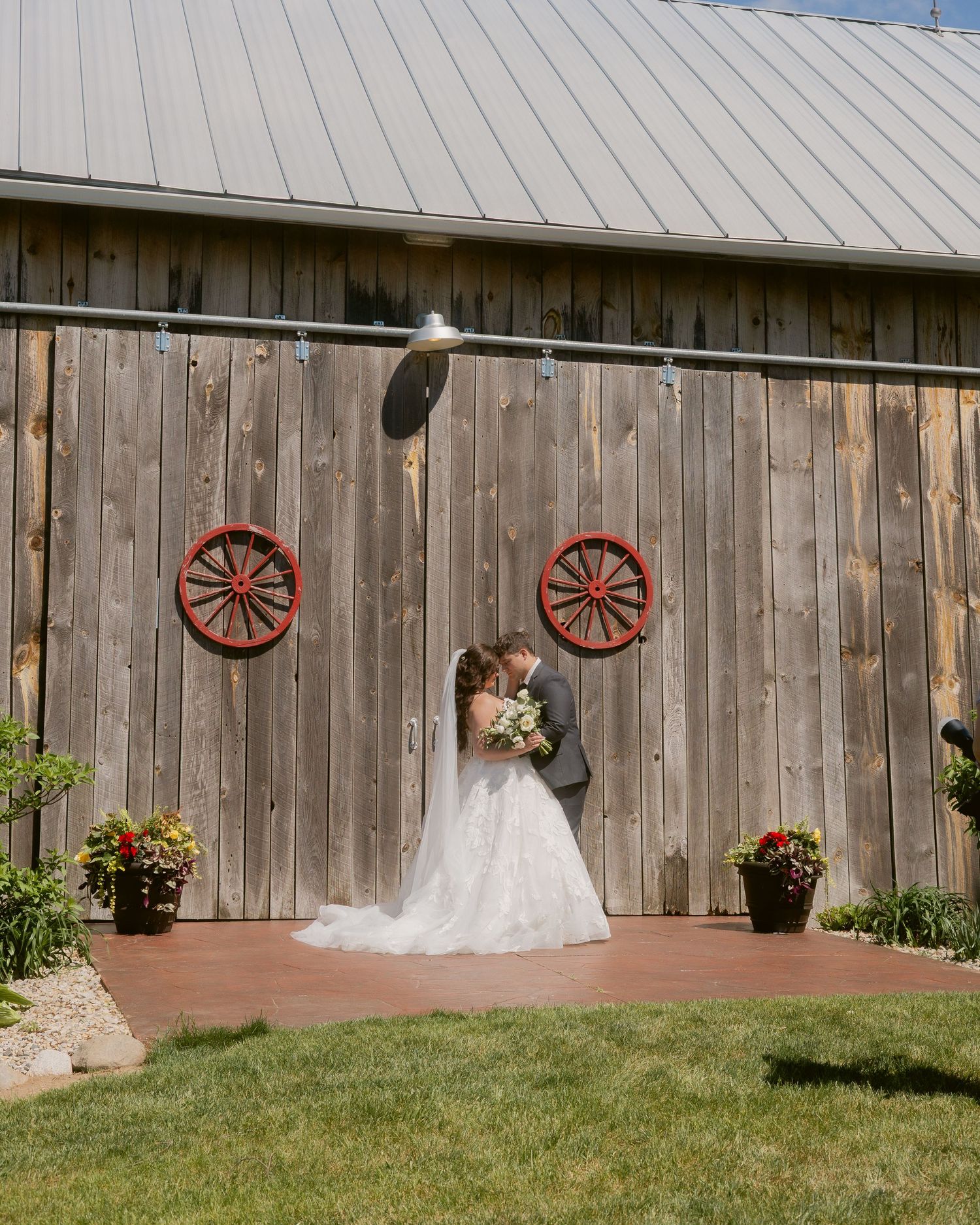 Newlyweds share a kiss in front of weathered barn doors adorned with red wagon wheel decor.