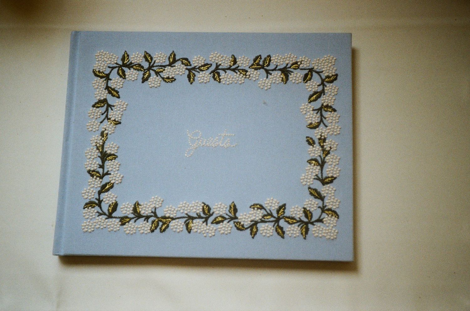 Vintage light blue wedding guest book with embroidered floral border and gold script on white wall.