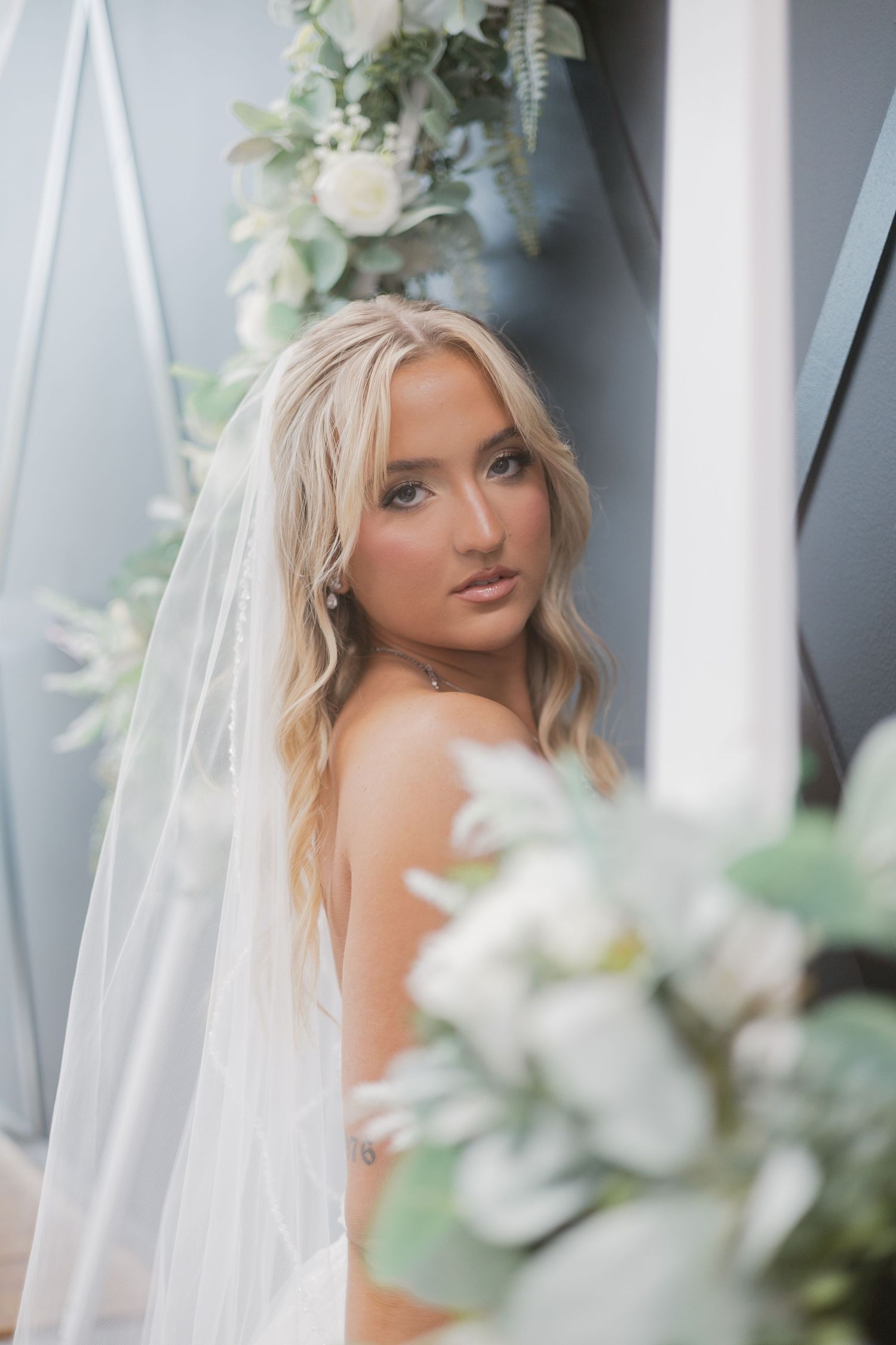 Stunning blonde bride in a white veil posing near floral arch with white roses and greenery.