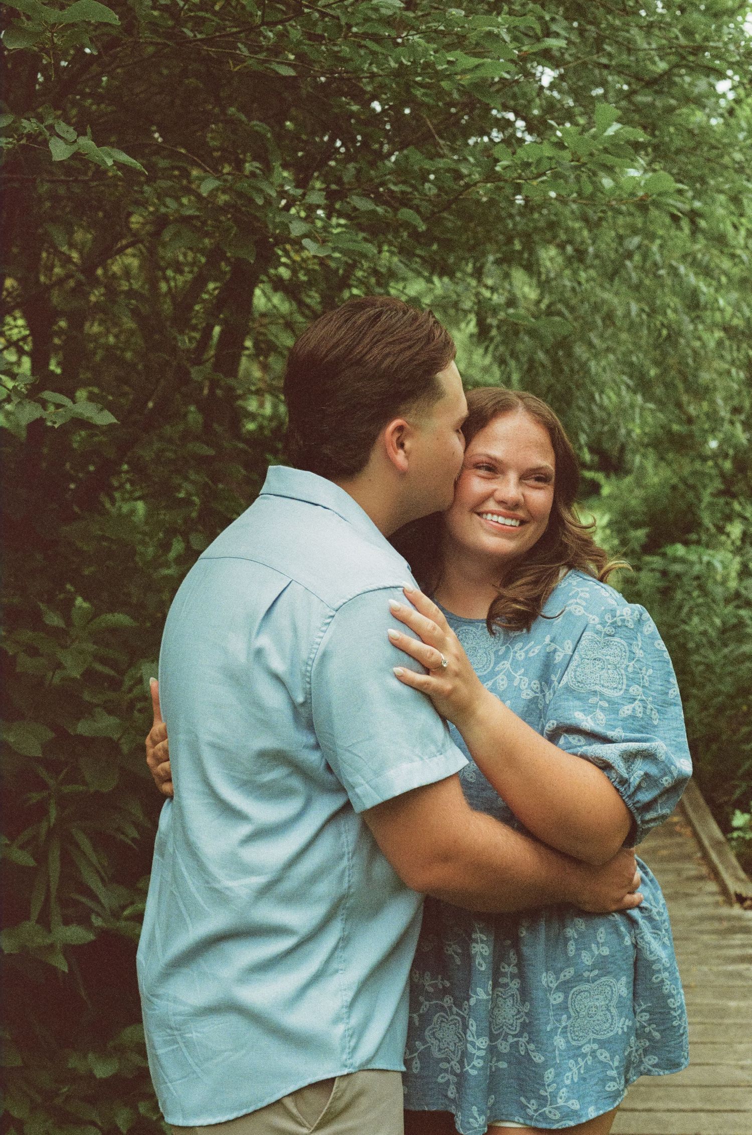 Couple embracing outdoors, man kissing woman's cheek while she smiles, surrounded by lush green trees.