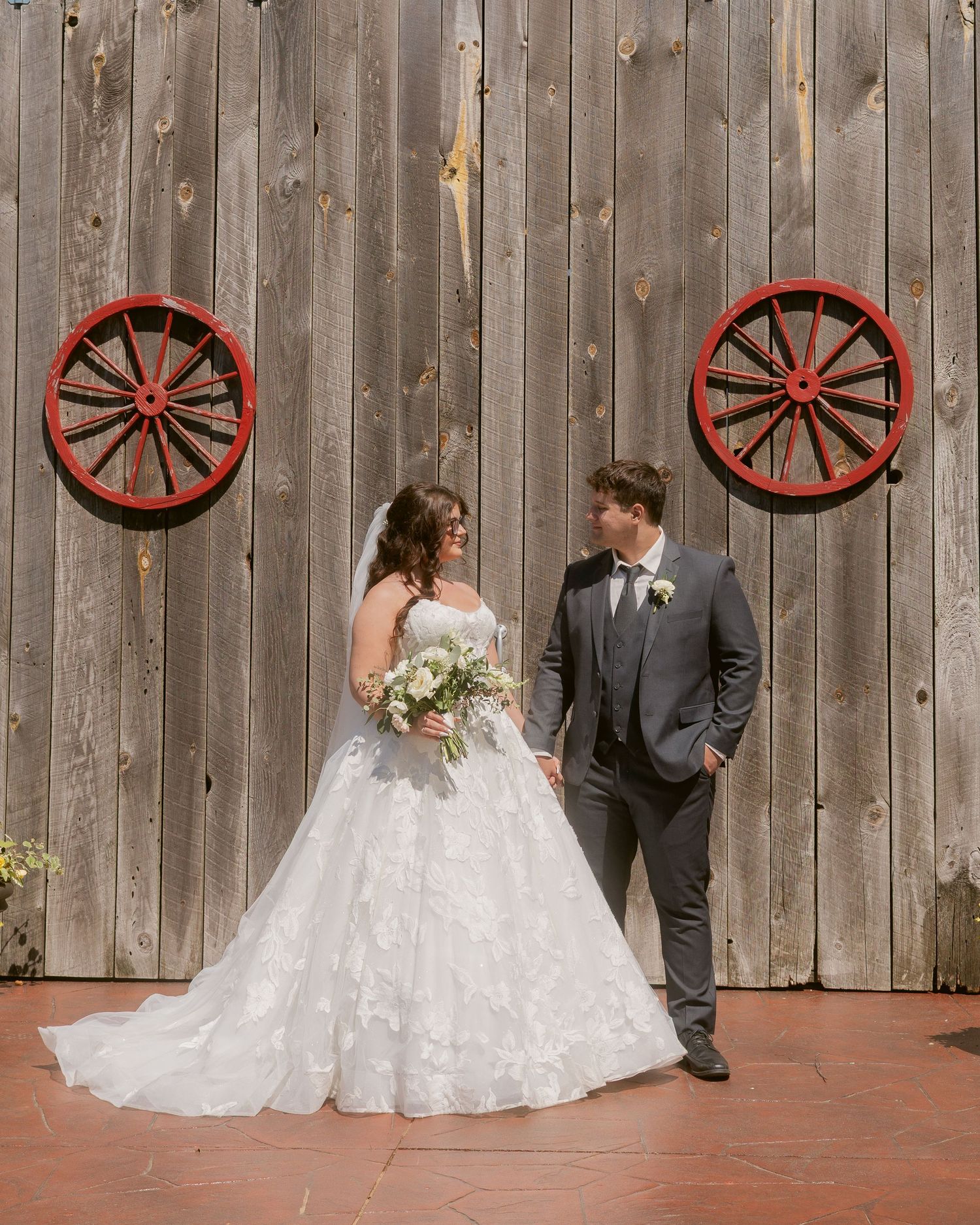 Bride and groom share loving glance before rustic barn wall decorated with red wagon wheels.