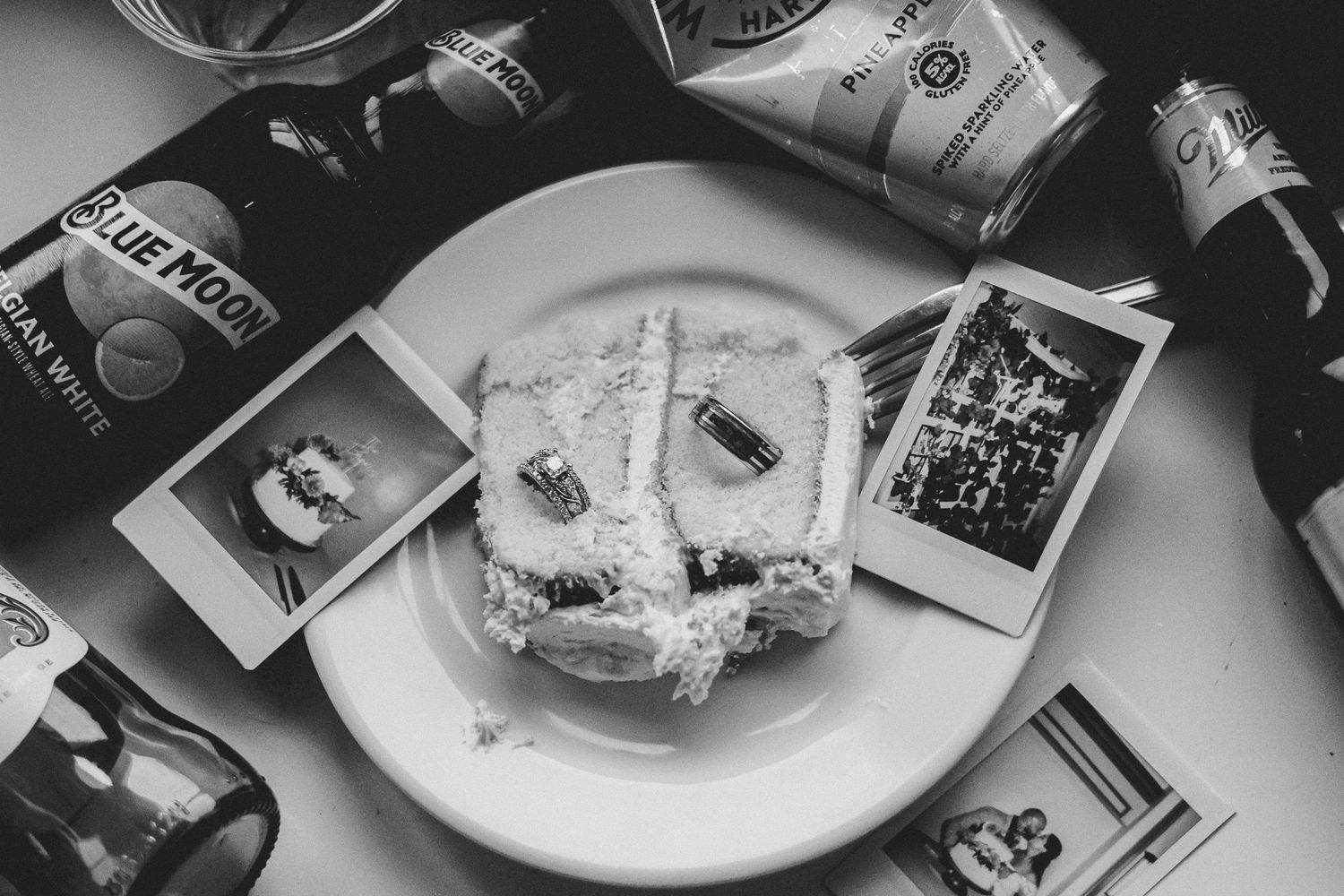 Black and white flat lay of wedding cake slice, Polaroid photos, rings, and beer bottles on plate.