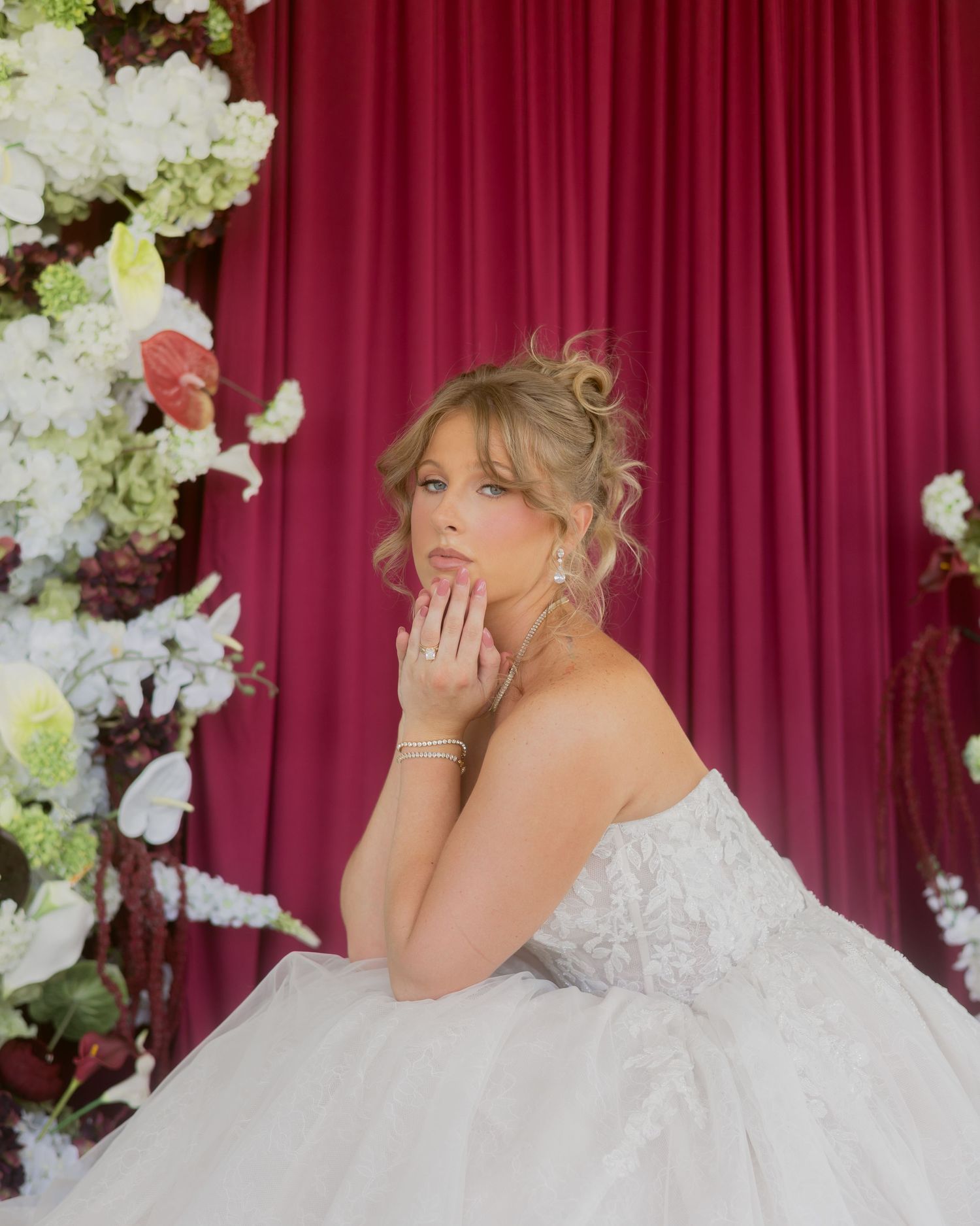 Bride in strapless lace gown posing thoughtfully beside lush white floral arrangements against a rich red velvet curtain.