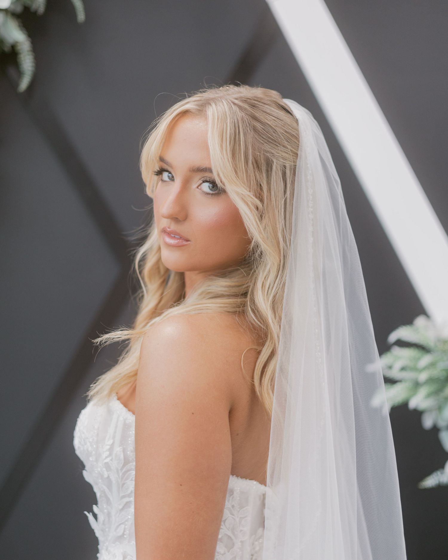 Blonde bride in a strapless lace wedding gown gazes over her shoulder while wearing a long flowing veil.