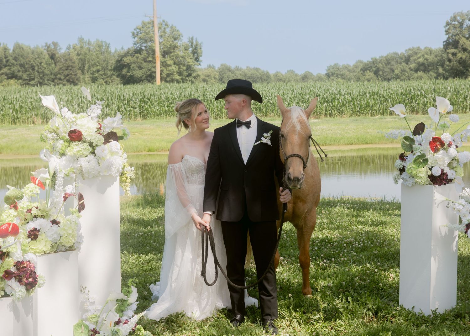 Bride and groom pose with a horse beside floral arrangements at an outdoor rustic wedding ceremony.
