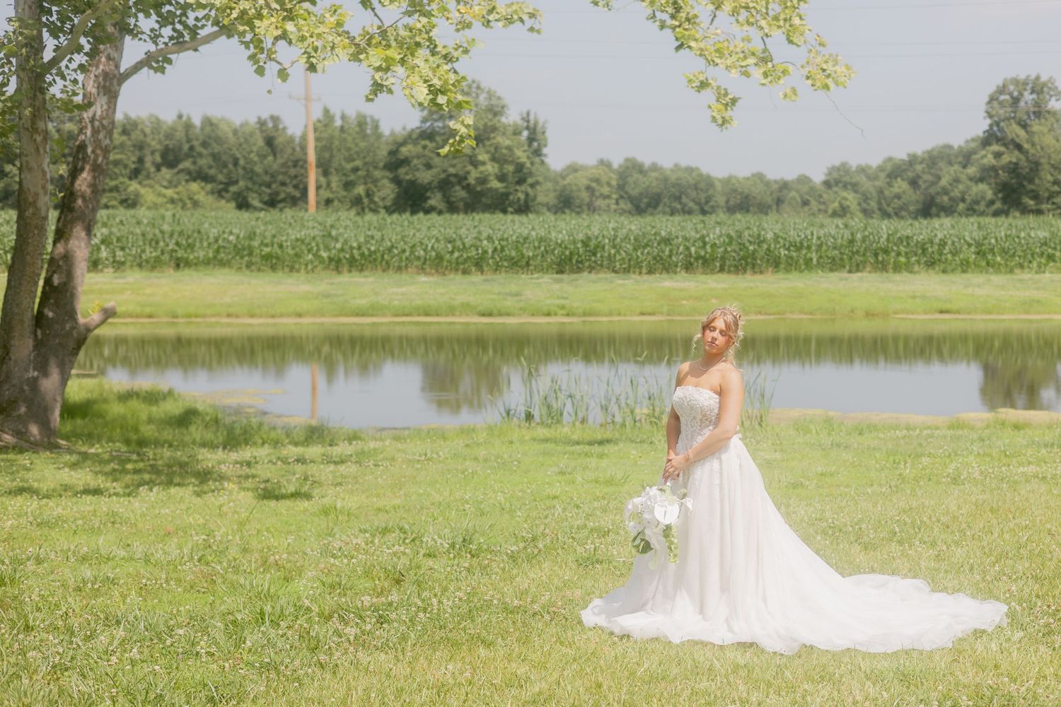 Beautiful bride in white strapless ballgown smiles while posing outdoors near a pond with lush green fields.