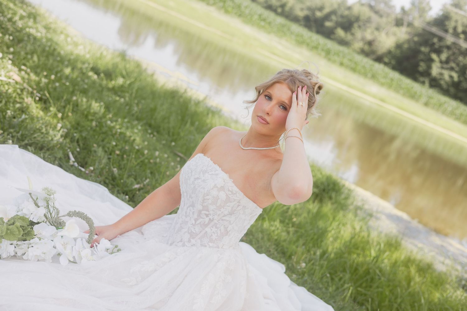 Bride in a strapless white wedding gown sitting by a peaceful lake in a green meadow.