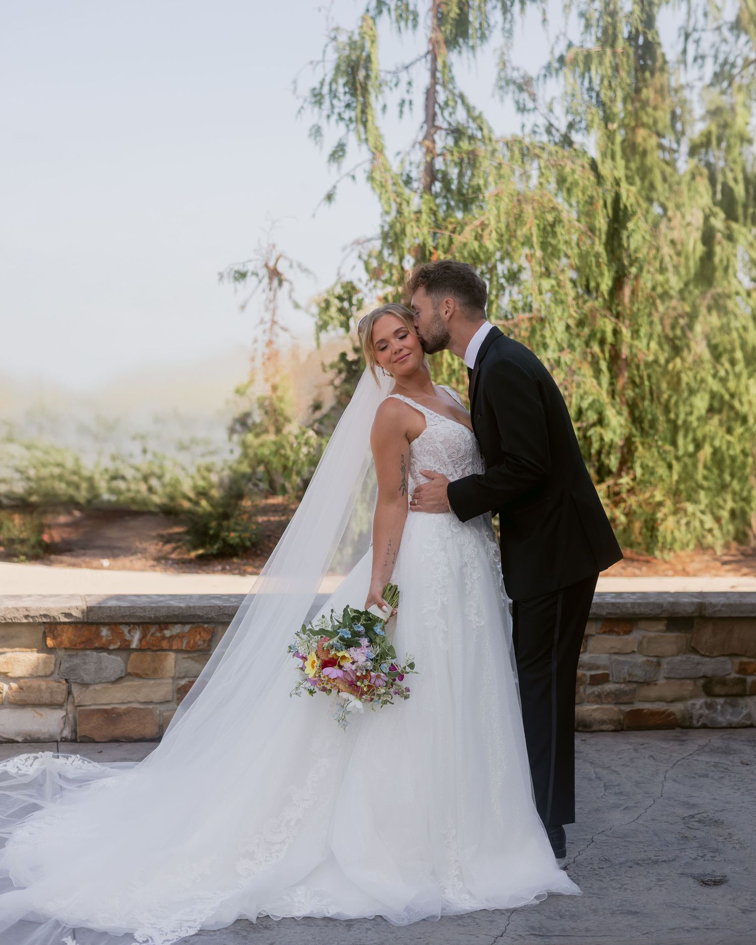 Bride and groom sharing romantic kiss outdoors with willow tree backdrop on their wedding day.