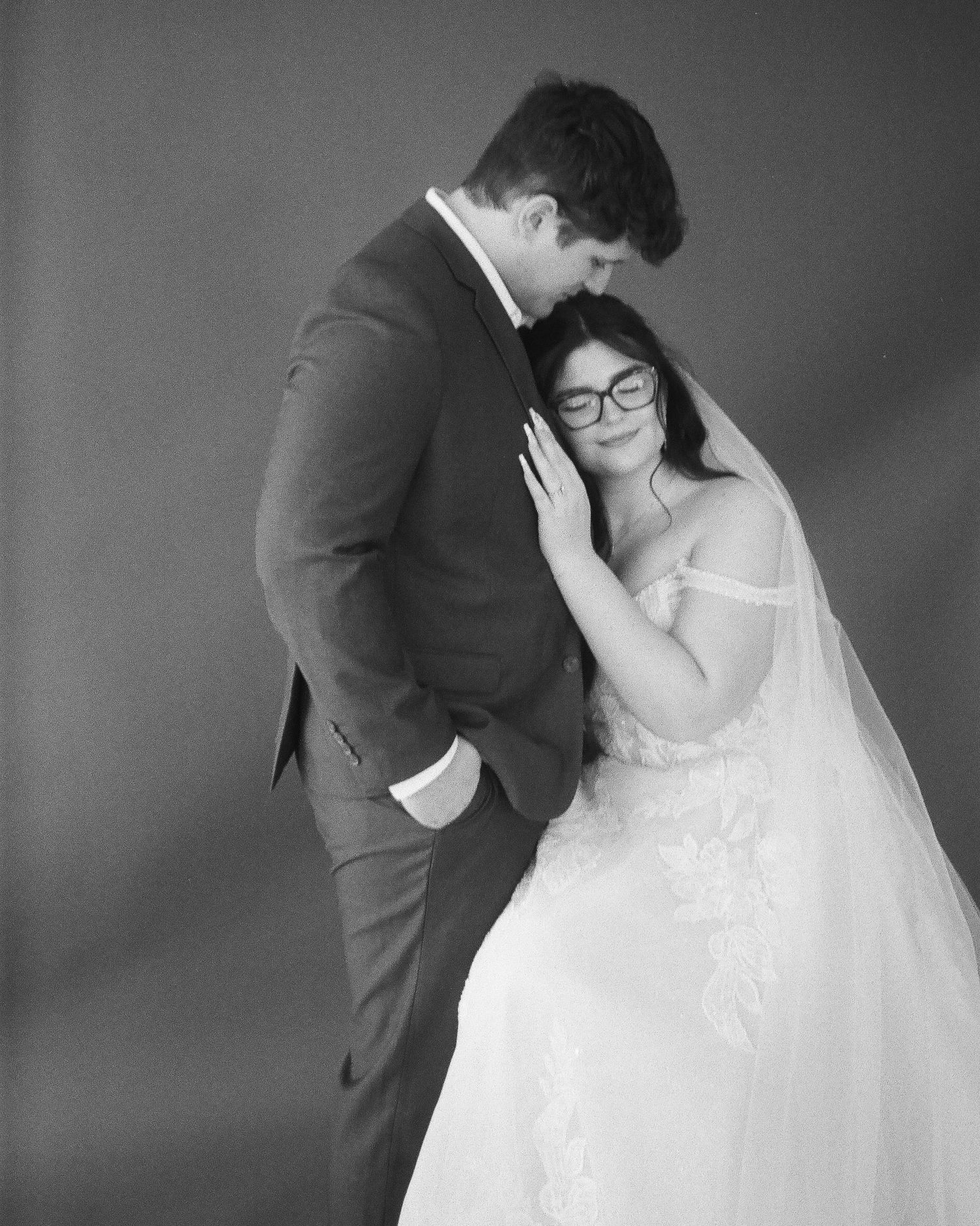 Intimate black and white portrait of a bride with glasses leaning against her groom in a studio setting.