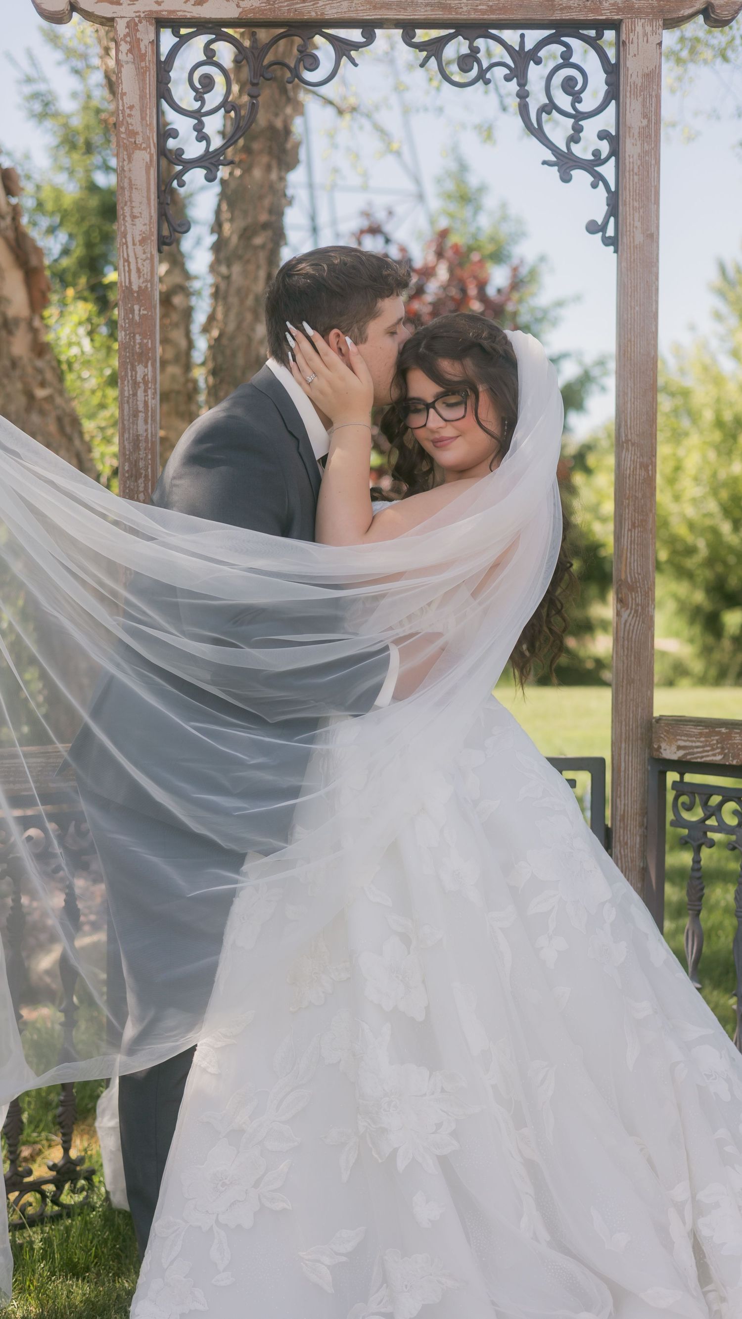 Bride and groom share romantic kiss under ornate wooden arch with veil flowing in the breeze.