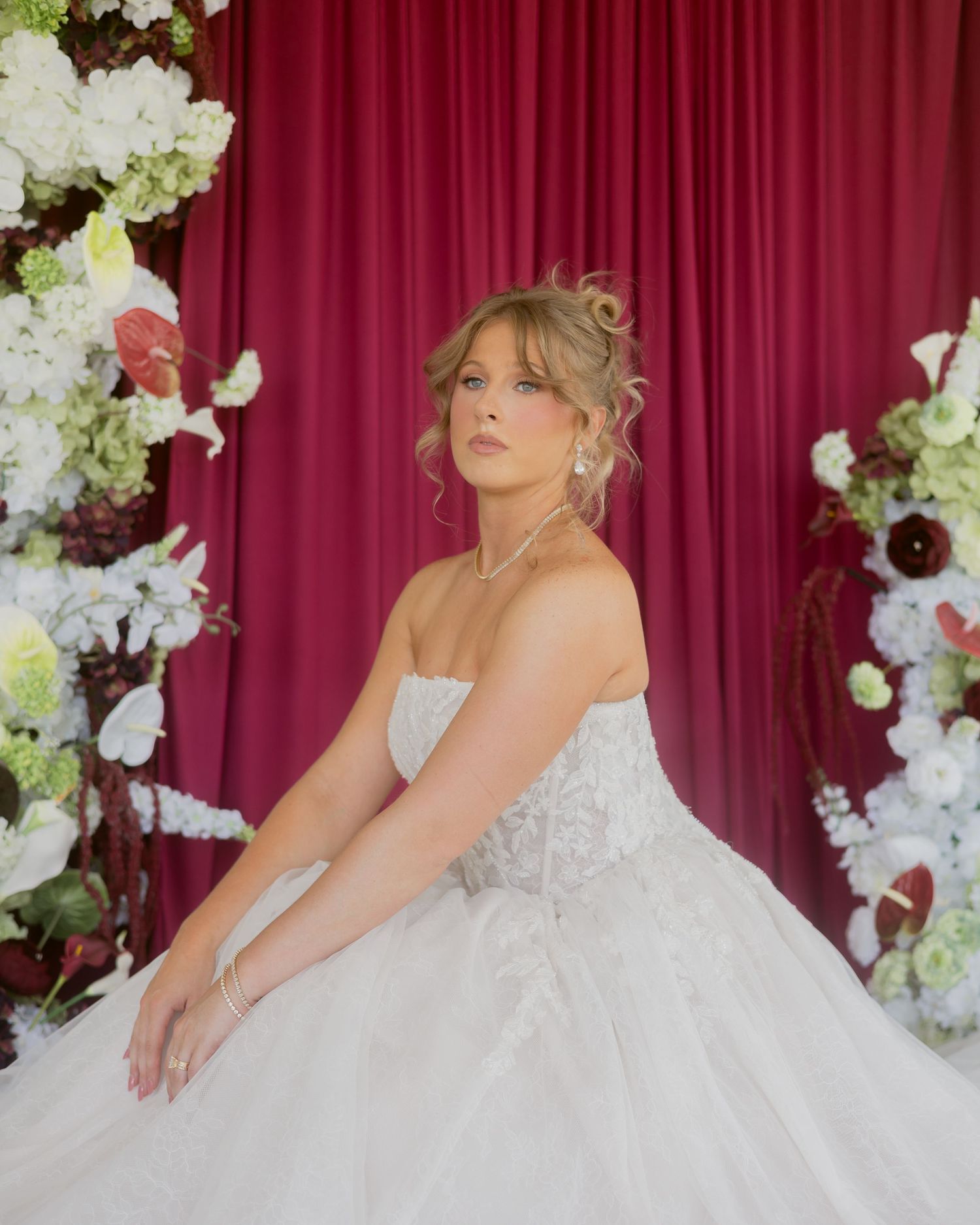 Elegant bride in strapless lace ball gown seated before a red velvet backdrop surrounded by white and burgundy florals.