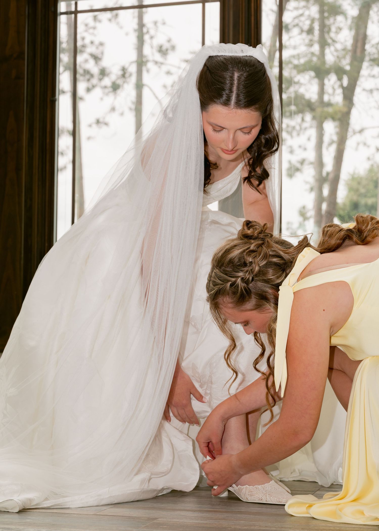 Bridesmaid in yellow dress helps bride with shoes while standing near large windows before ceremony.