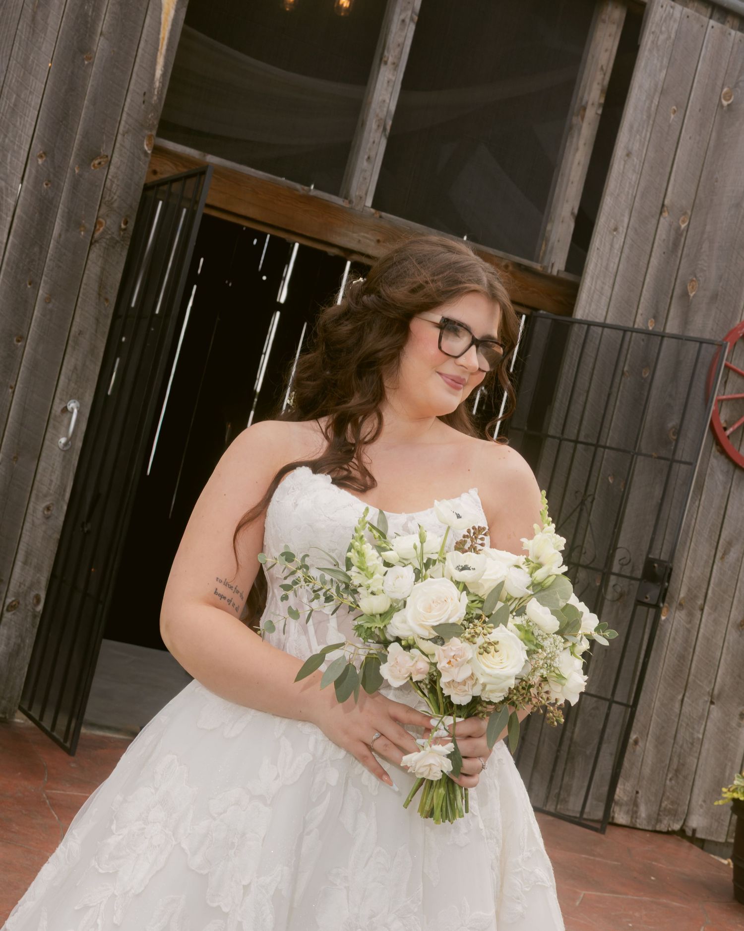 Bride in strapless lace wedding gown holding white floral bouquet, standing at rustic barn entrance.