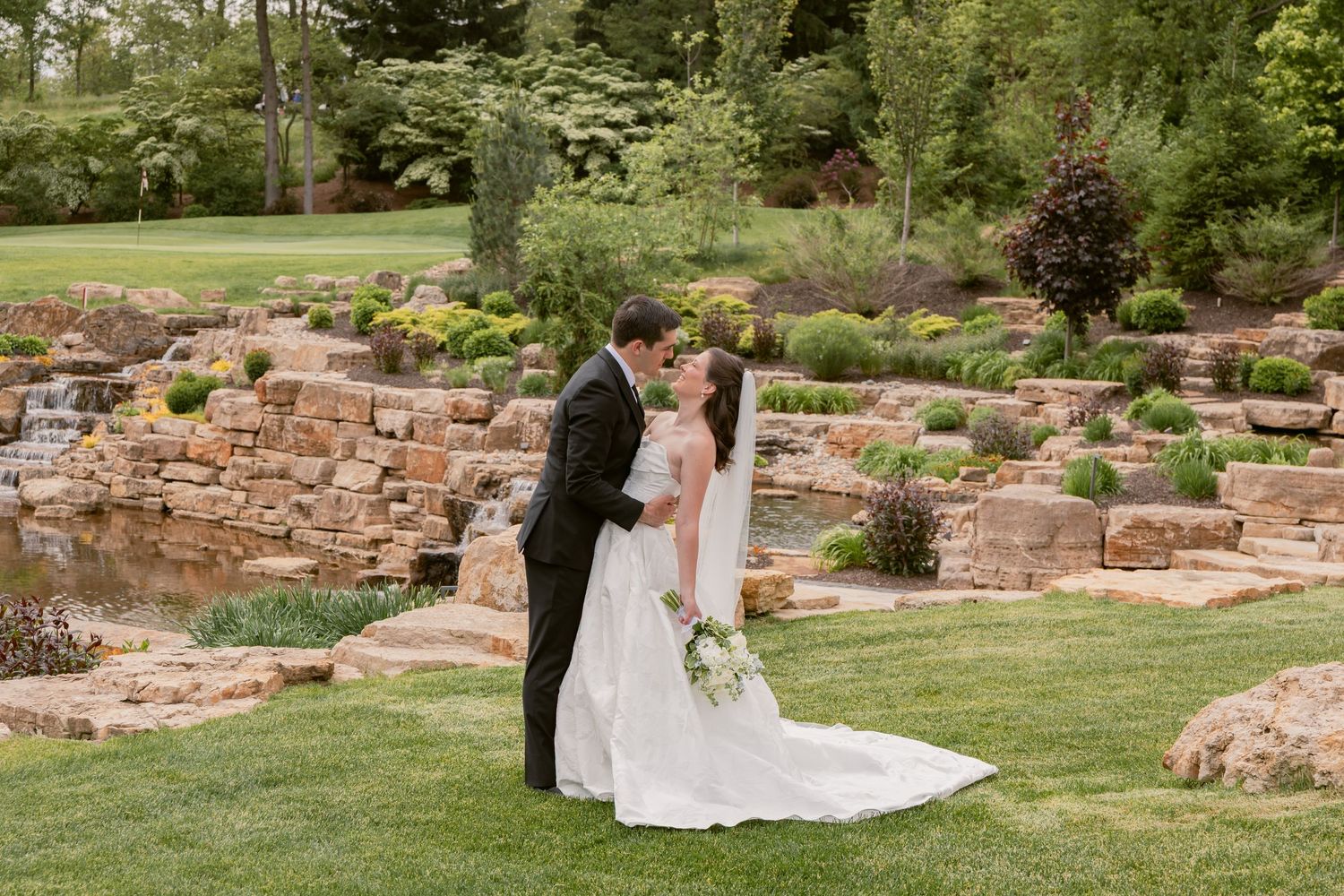 Bride and groom sharing a kiss near a stone waterfall garden, with lush greenery surrounding them.
