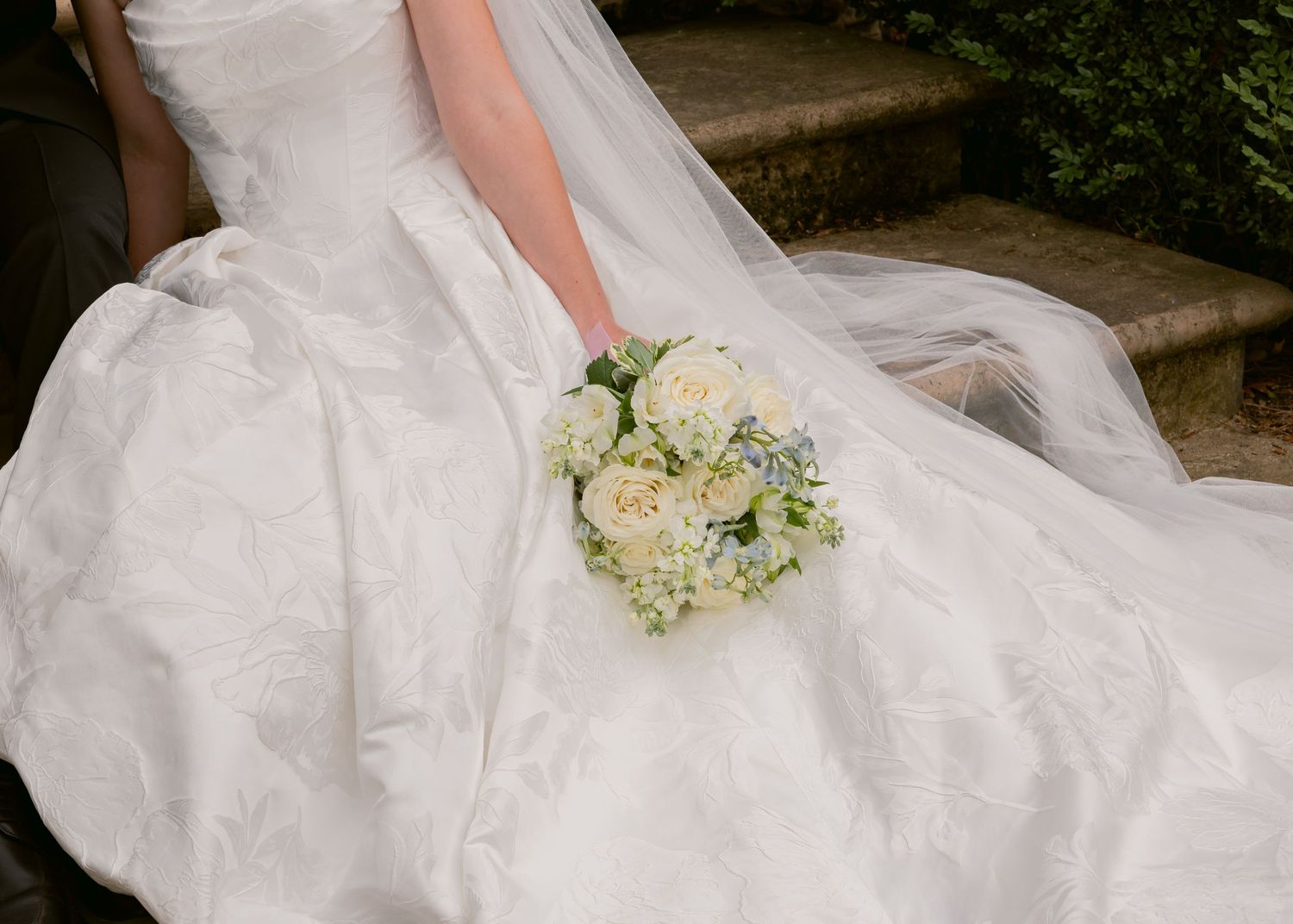 Close-up of a bride in a white ball gown holding a classic white rose and greenery wedding bouquet.