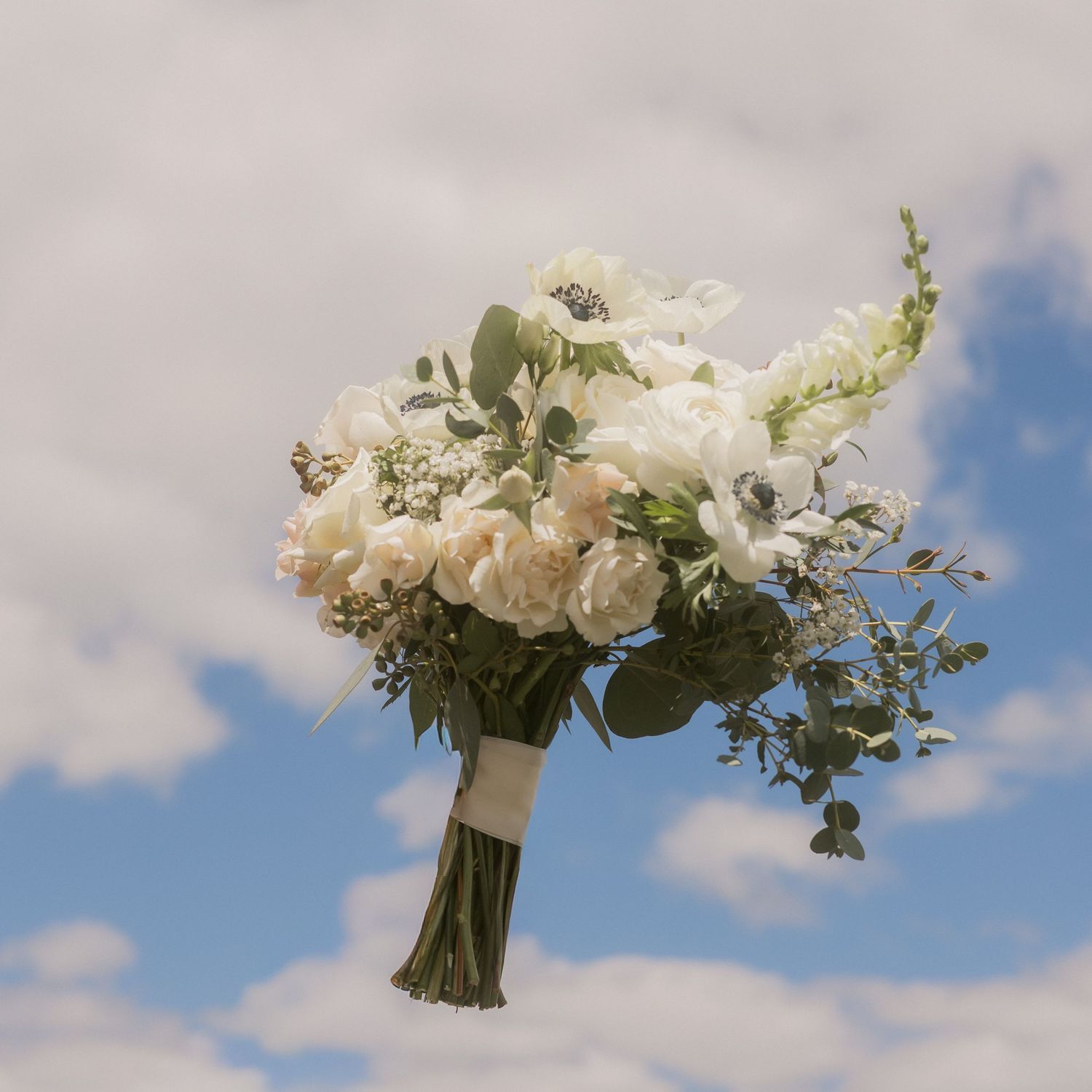 Elegant white wedding bouquet with anemones and roses held against a bright blue cloudy sky.
