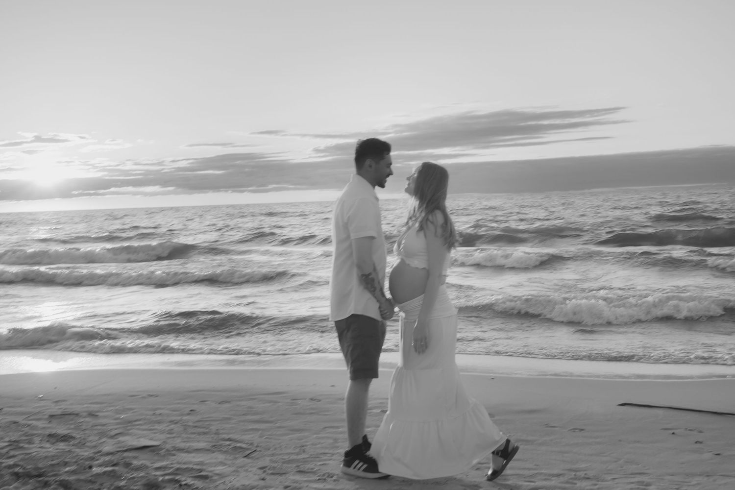 Black and white photo of pregnant couple holding hands on beach at sunset, ocean waves in background.