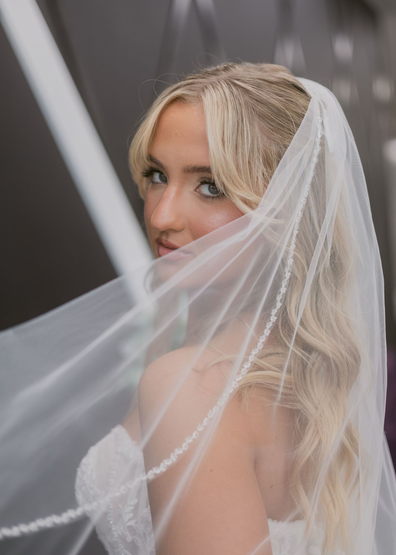 Bride playfully peeks through her pearl-trimmed veil with a soft smile in a stunning close-up portrait.
