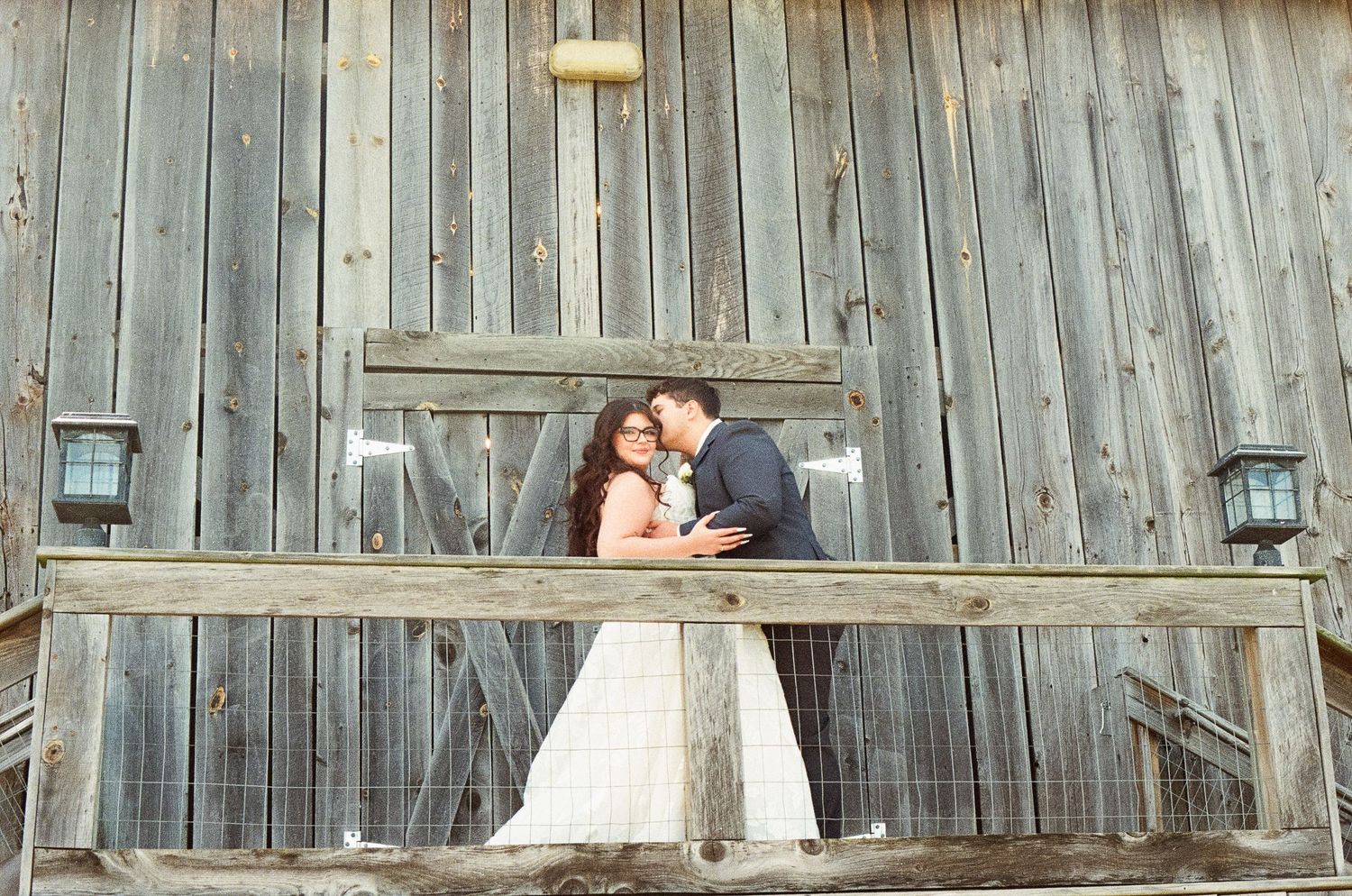 Bride and groom share a tender kiss on rustic barn balcony with weathered wooden backdrop.
