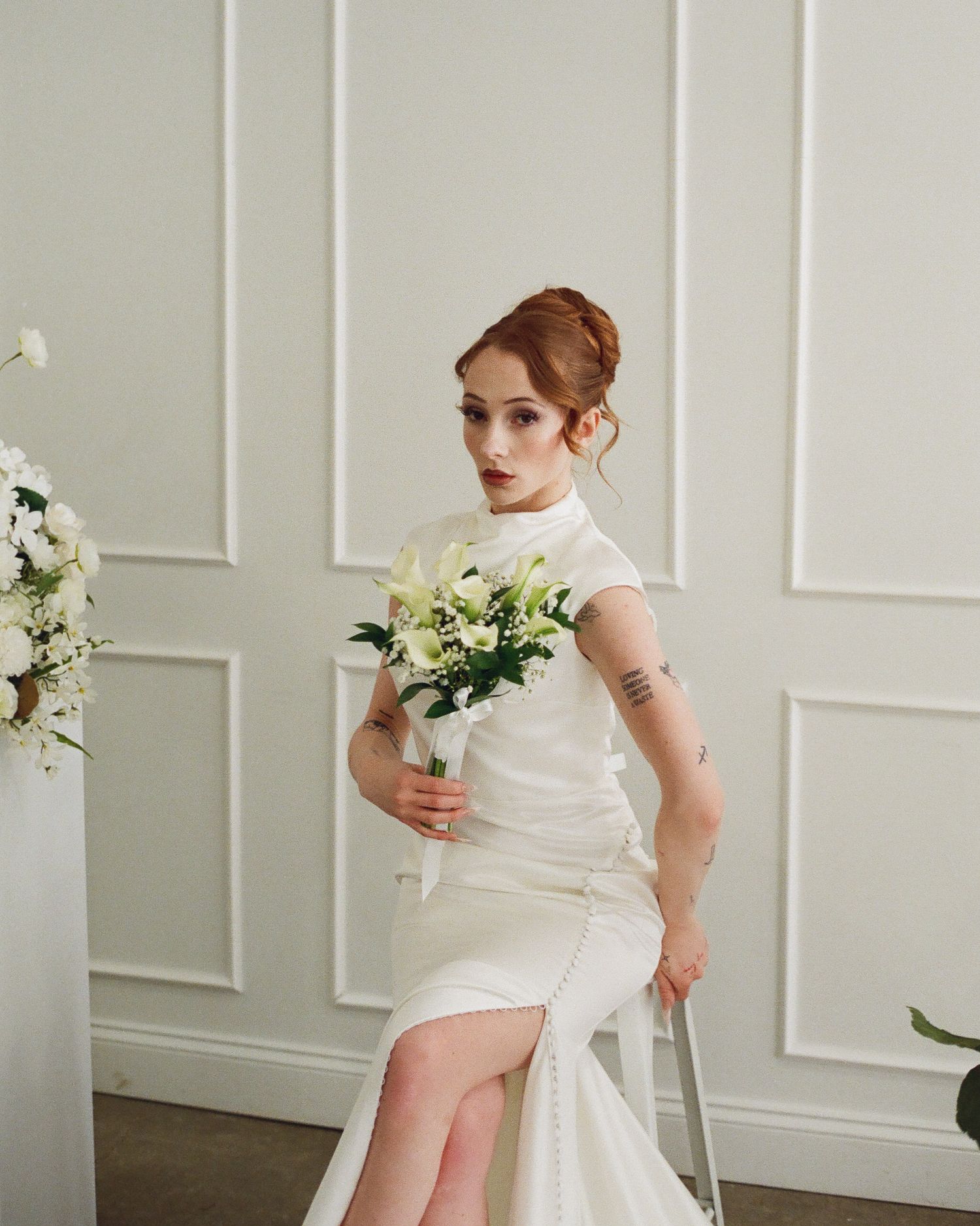 Red-haired bride in sleek white dress holds white lily bouquet beside floral arrangement indoors.