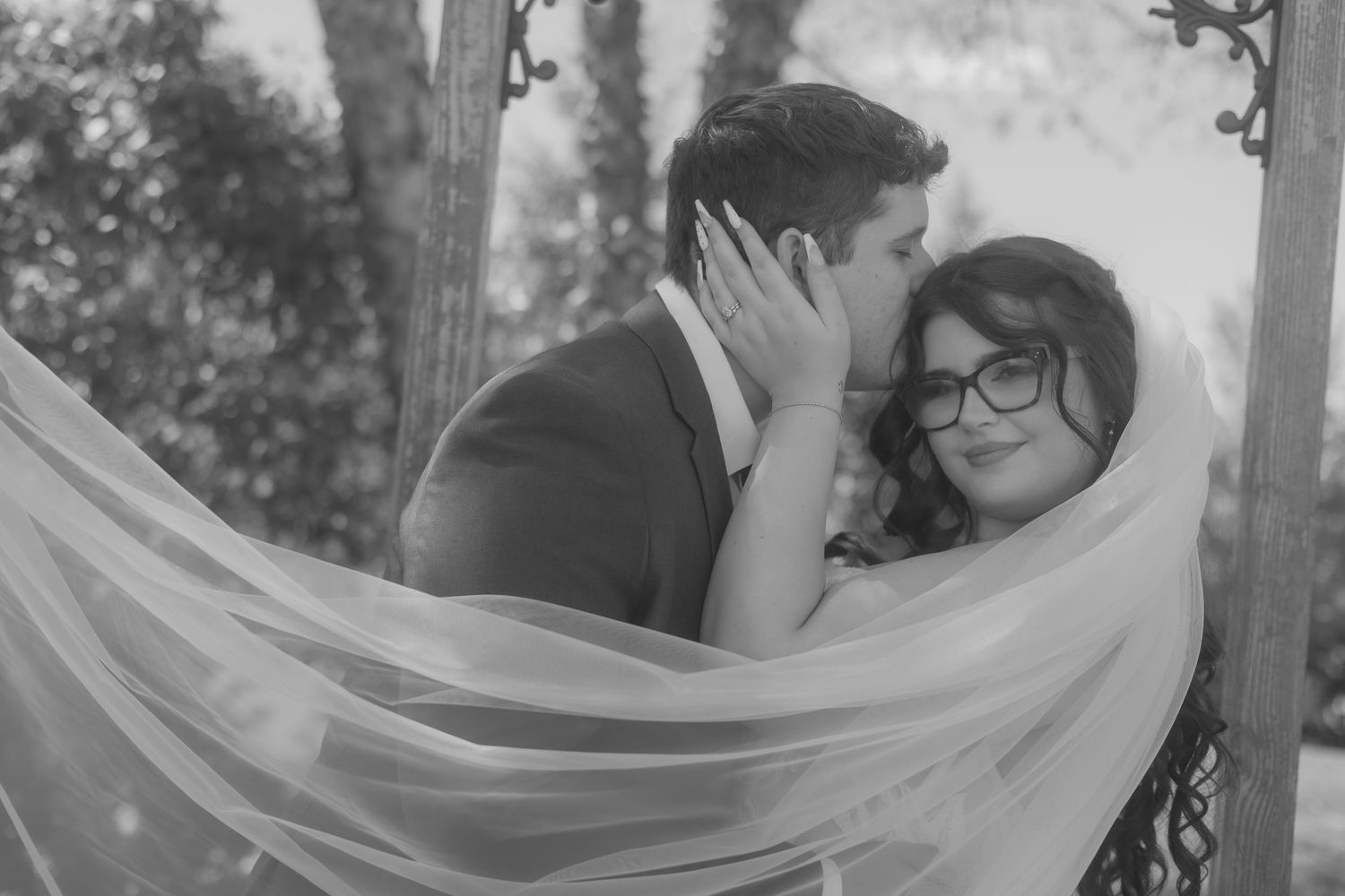 Black and white photo of a couple sharing a romantic kiss under a floral arch on their wedding day.